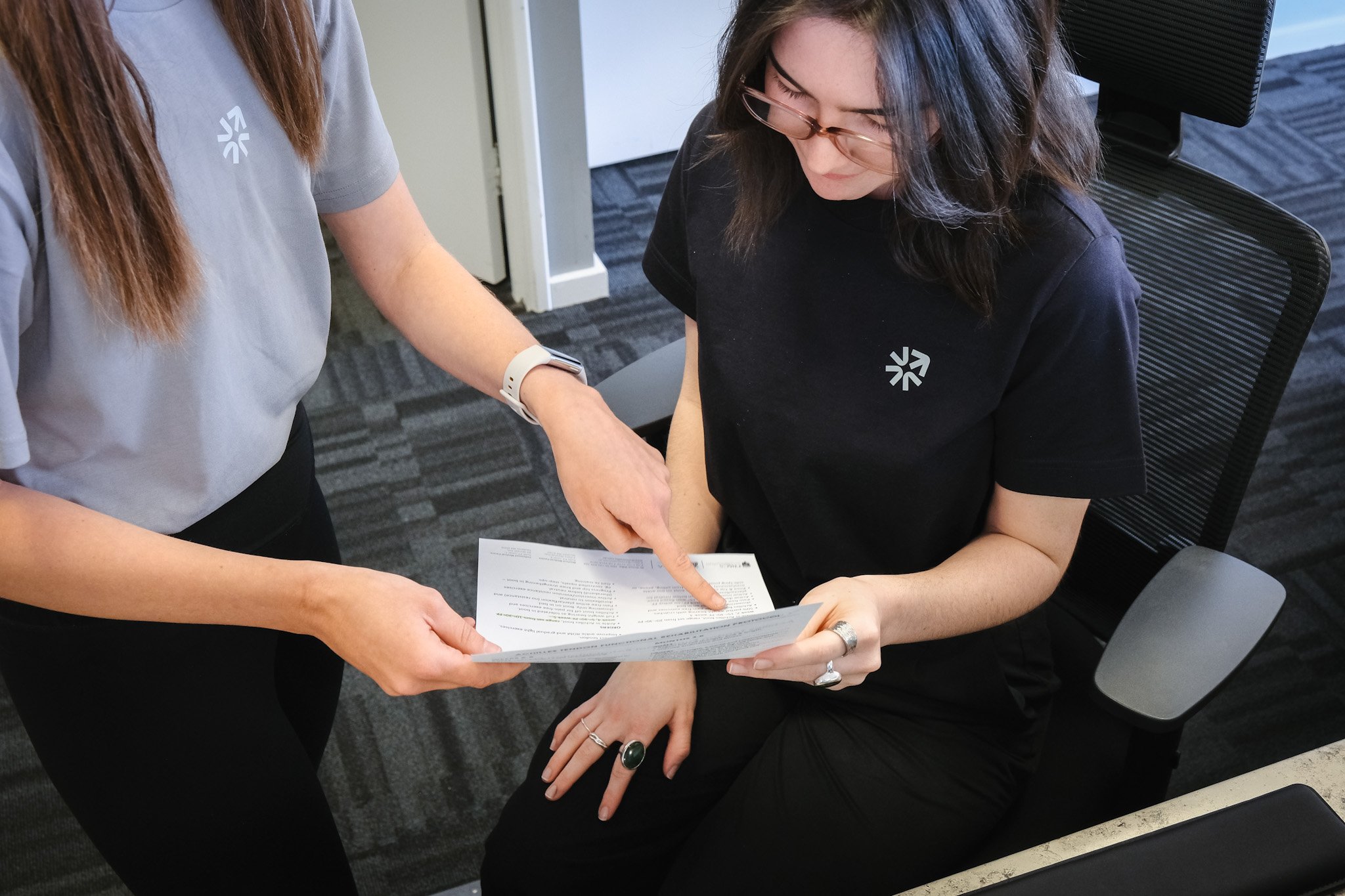 Bunbury Physio - Two women are examining a document together in an office setting. One woman is standing, wearing a gray shirt, and the other is sitting, wearing a black shirt and glasses, pointing at the paper.