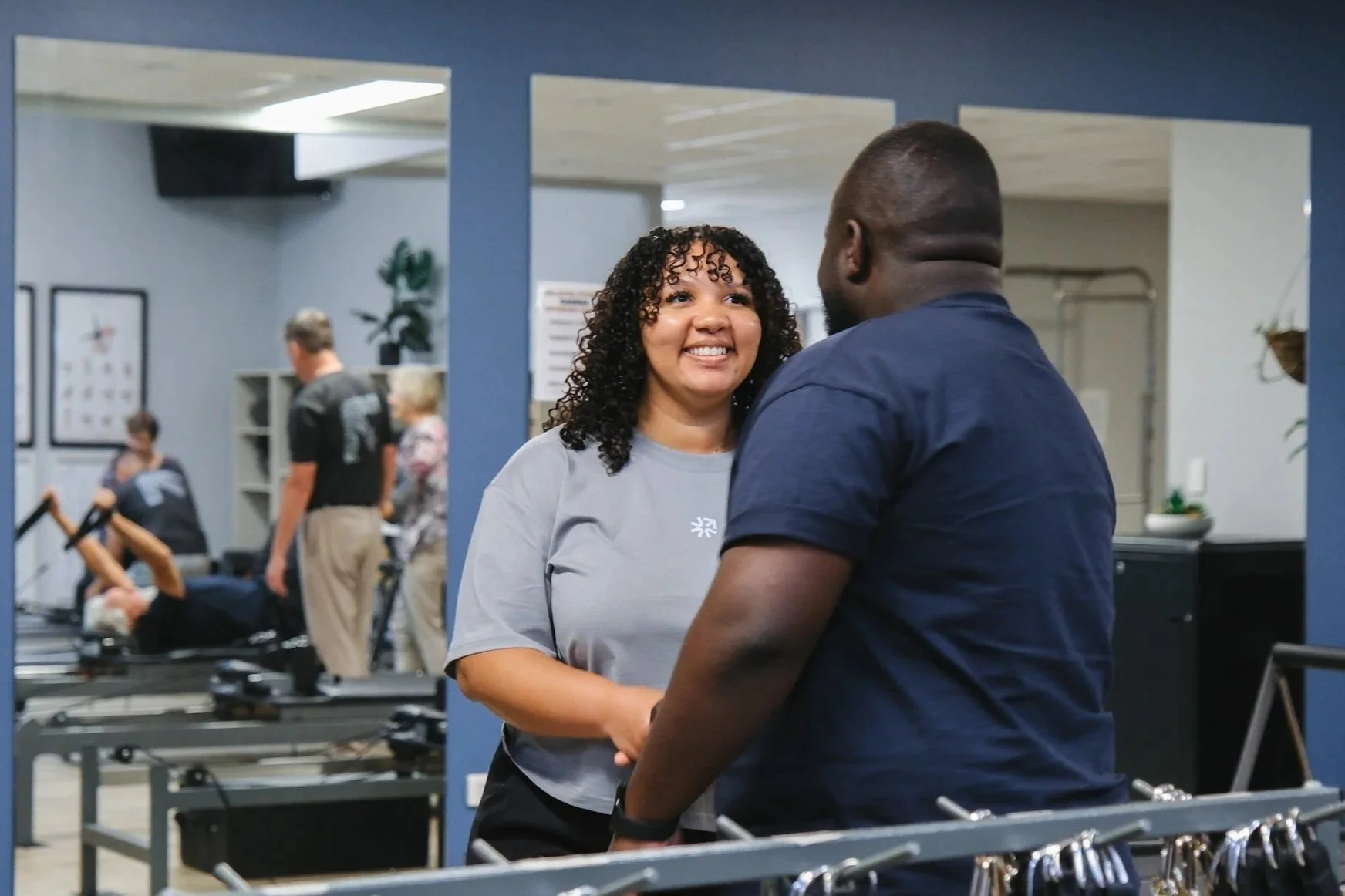 Bunbury Physio - A woman and a man are smiling and holding hands, standing in a gym or physical therapy center. There are other people working out or engaging in activities, gym equipment, and decor.