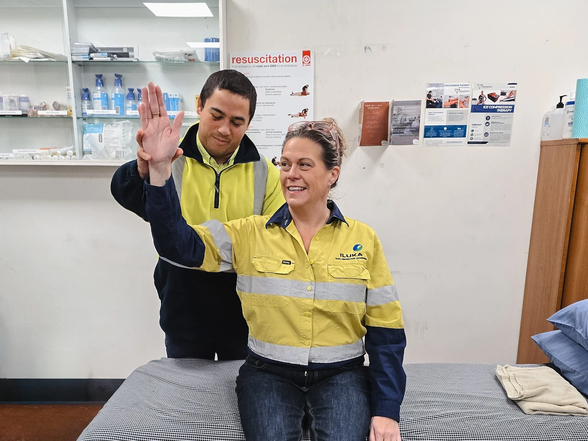 Two emergency medical responders, a man and a woman, are performing a medical procedure or demonstration. The man is in a yellow and black uniform and is raising his hand, while the woman, also in a uniform, is sitting with her eyes closed. They are in a medical setting with supplies and informational posters in the background.
