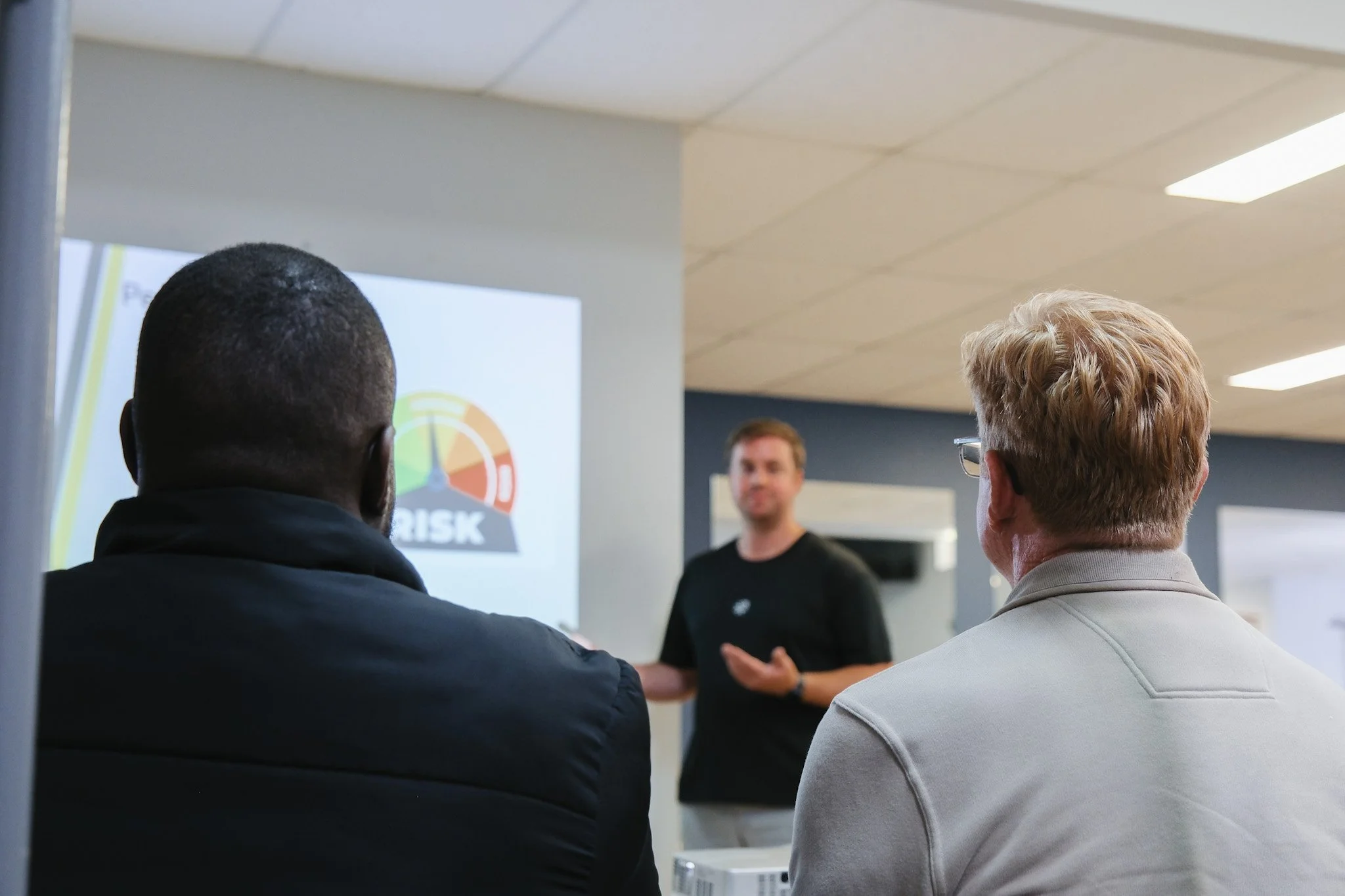 Bunbury Physio - Two men are listening to a man giving a presentation in a conference room. A chart with a risk gauge is displayed on a screen behind the presenter.