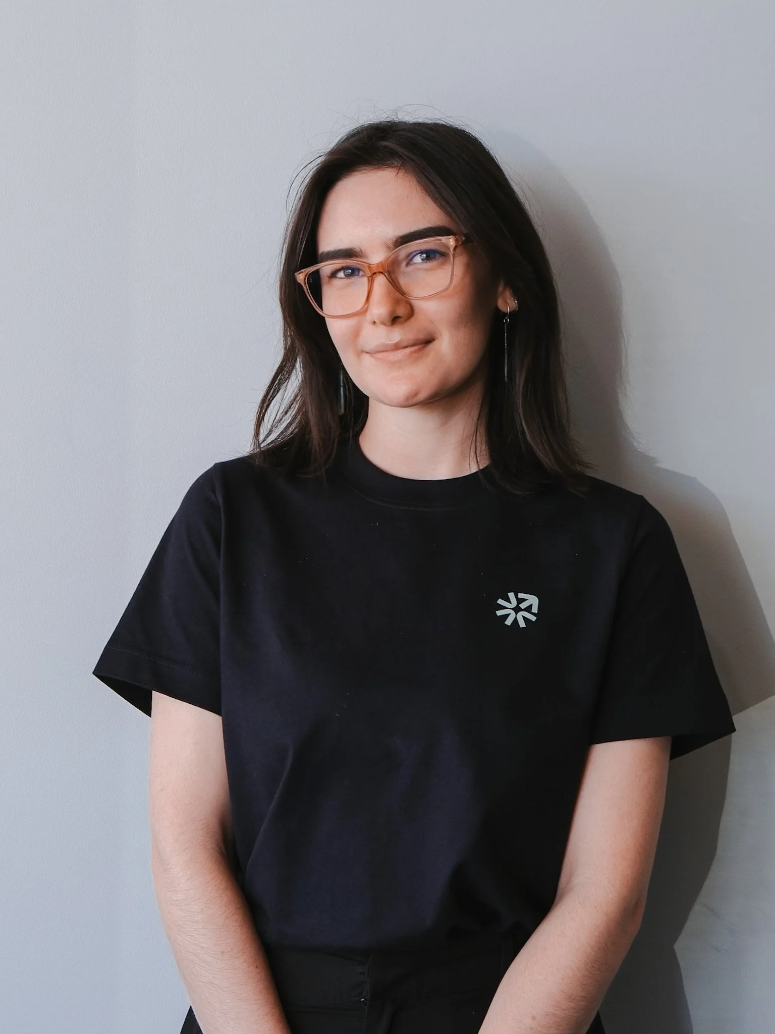 Bunbury Physio - Close-up of a young woman with dark hair and glasses, wearing a black shirt, standing against a plain white wall.