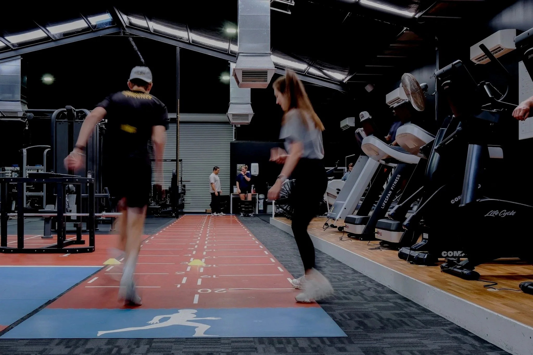Bunbury Physio - People exercising on a treadmill in a gym with a running track in the foreground and workout equipment along the walls.