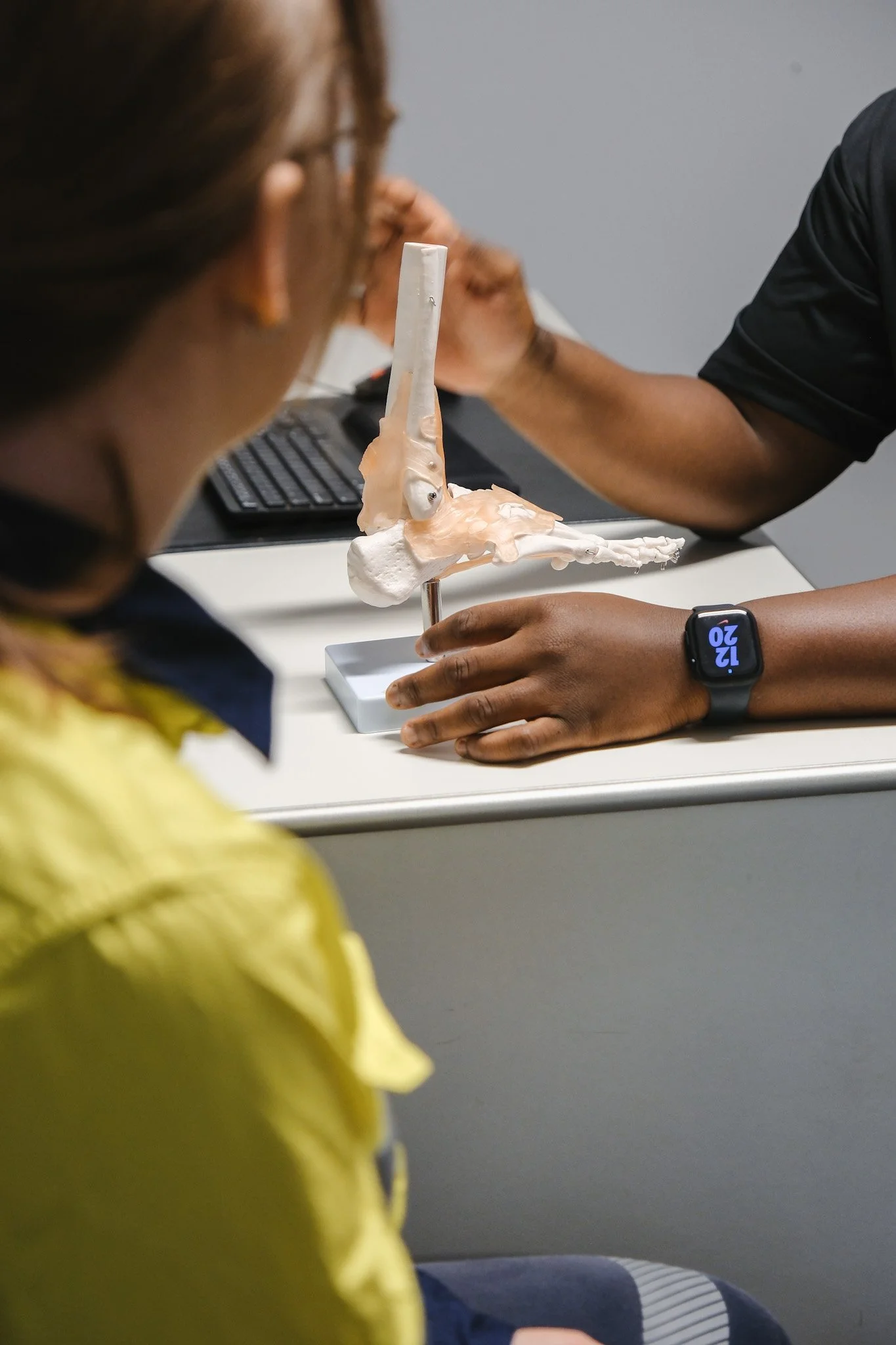 Bunbury Physio - A person is holding a model of a human pelvis and spine, with another individual sitting nearby, in an office setting with a computer keyboard and monitor.