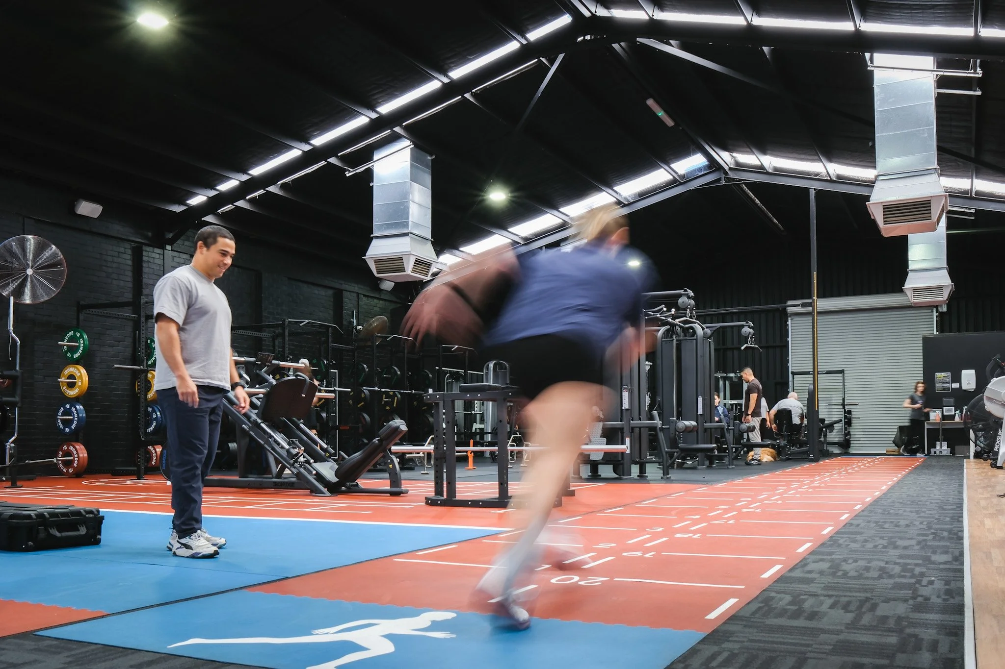 Bunbury Physio - People working out in a gym, with one person standing and others in motion doing exercises, gym equipment visible in the background, and a black ceiling with skylights.