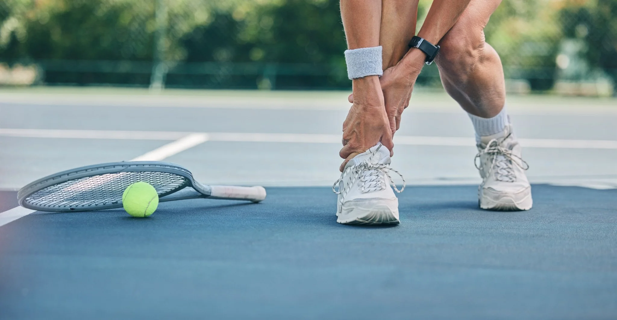 Bunbury Physio - A person on a tennis court bending down clutching their ankle, experiencing a sports injury, with a tennis racket and tennis ball lying on the court nearby.