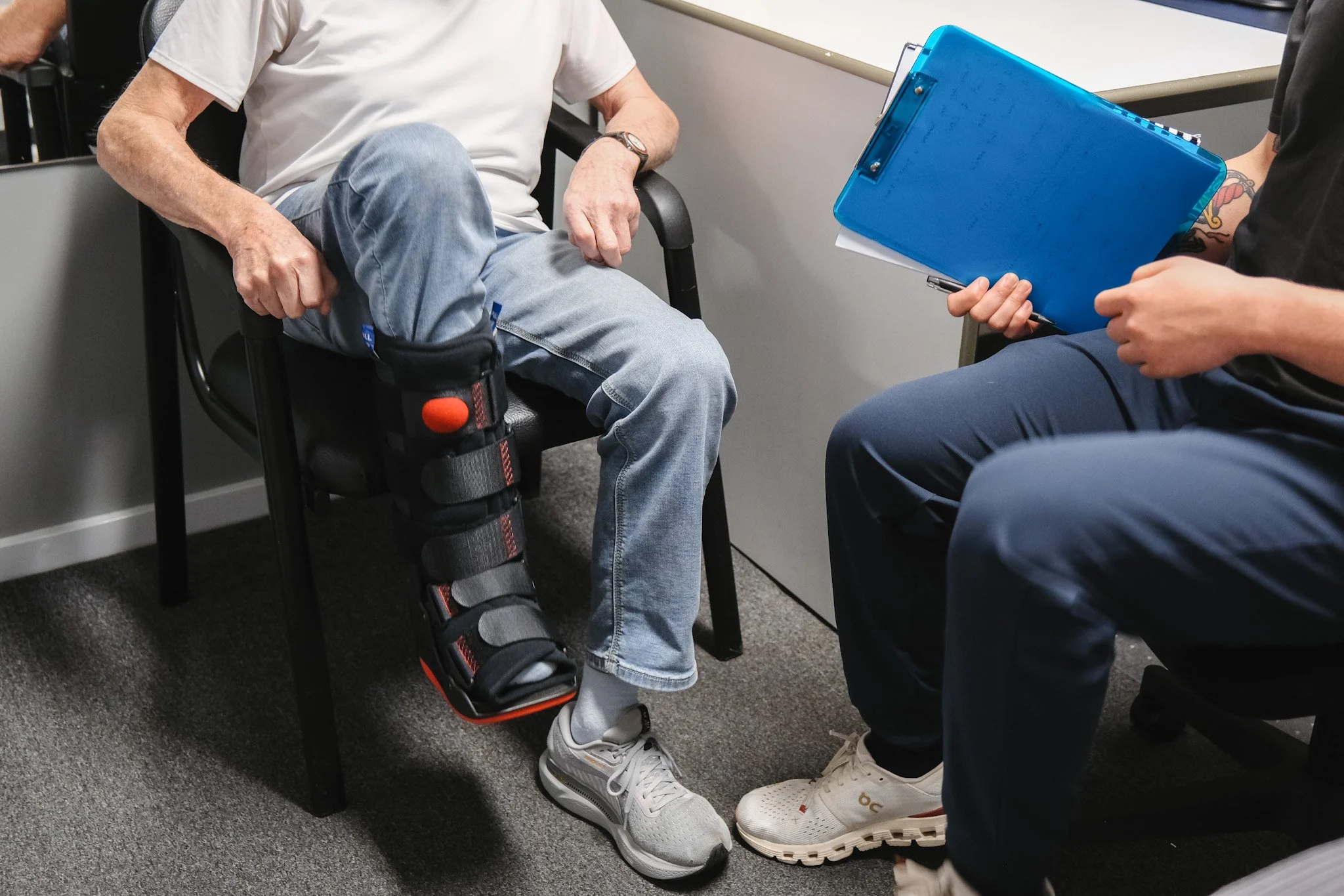 Bunbury Physio - Person with a medical leg brace sitting in a chair, having a conversation with another person holding a clipboard in a clinical setting.