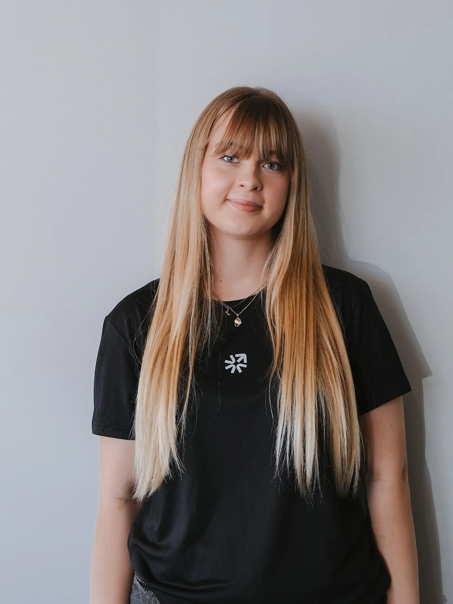 Bunbury Physio - A young woman with long straight blonde hair and bangs standing against a plain light gray wall, smiling softly at the camera.