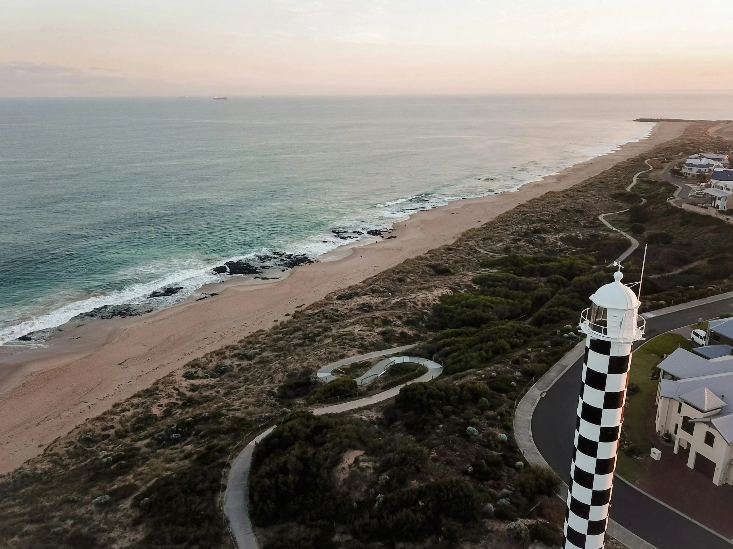 Bunbury Physio - View of a beach with dunes and a winding pathway, residential houses along the coast, and a lighthouse with a black and white checkerboard pattern in the foreground. Ships are visible on the horizon in the ocean.