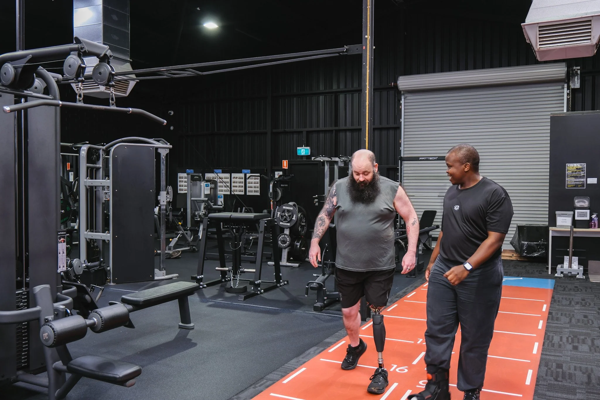 Bunbury Physio - Two men are in a gym, one with a prosthetic leg. They are walking on a marked track on the floor, surrounded by gym equipment and black industrial walls.
