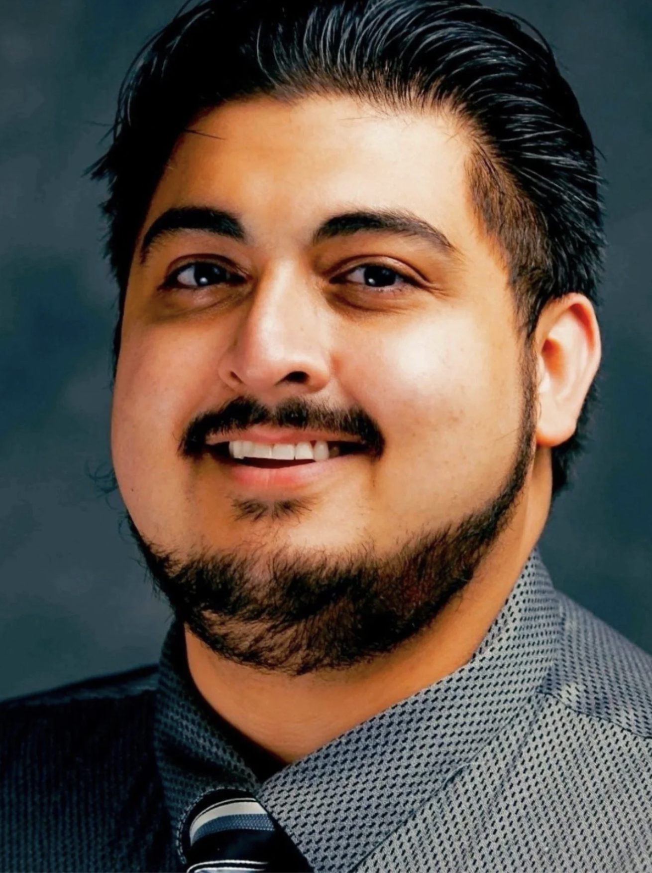 A close-up portrait of a smiling man with dark hair, a mustache, and a beard, wearing a patterned shirt and a tie, against a dark background.