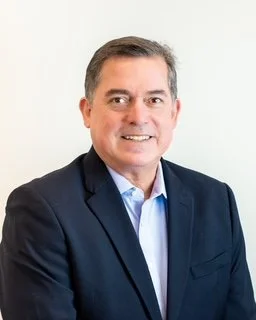 A middle-aged man in a dark suit and light blue shirt smiling, standing against a plain off-white background.