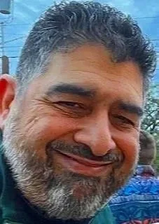 Close-up of a smiling man with dark, gray, and black hair and a gray beard, outdoors.