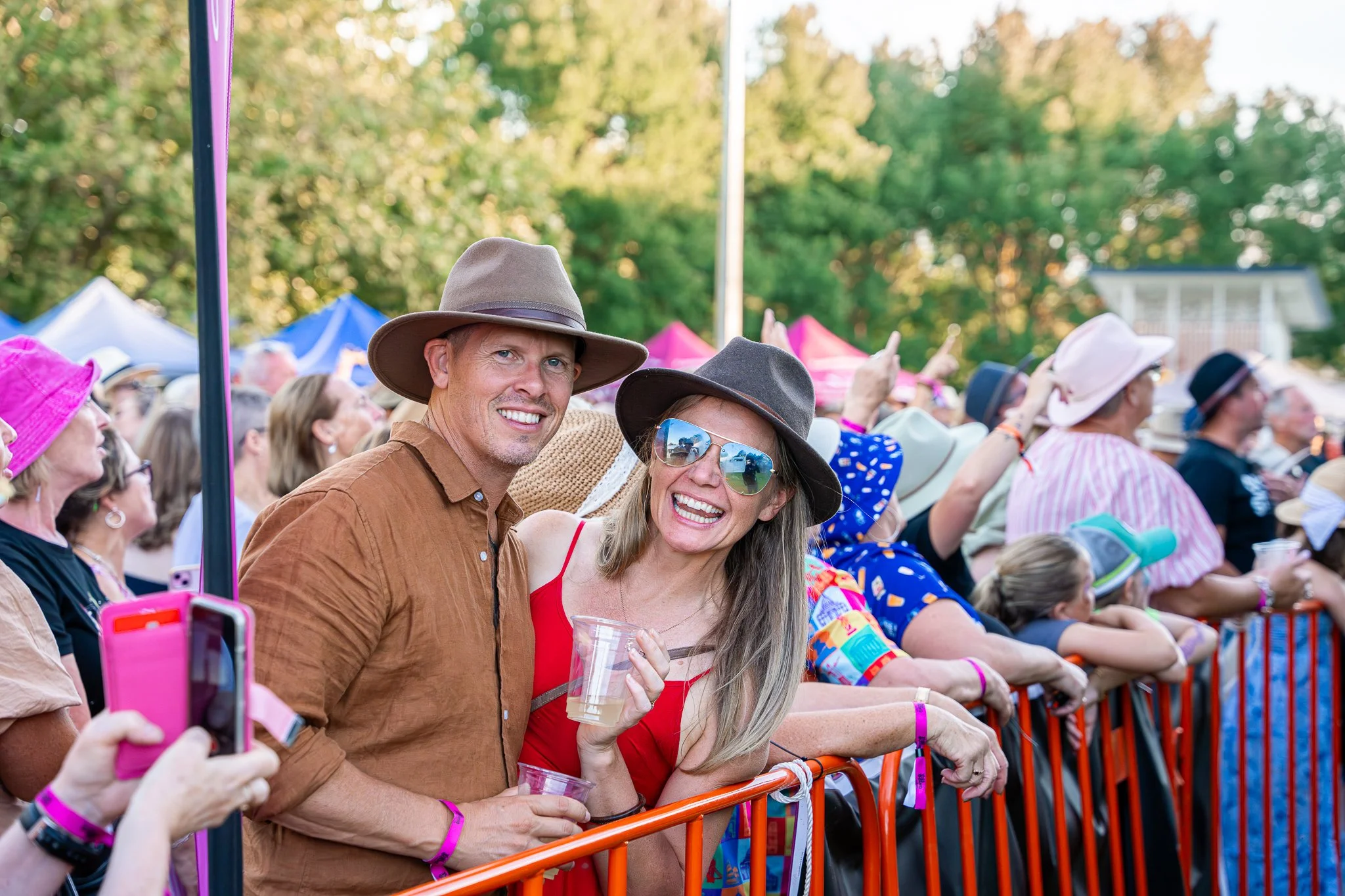Crowd of cheerful people at an outdoor event, wearing stylish hats, some holding drinks, with colorful tents and greenery in the background.