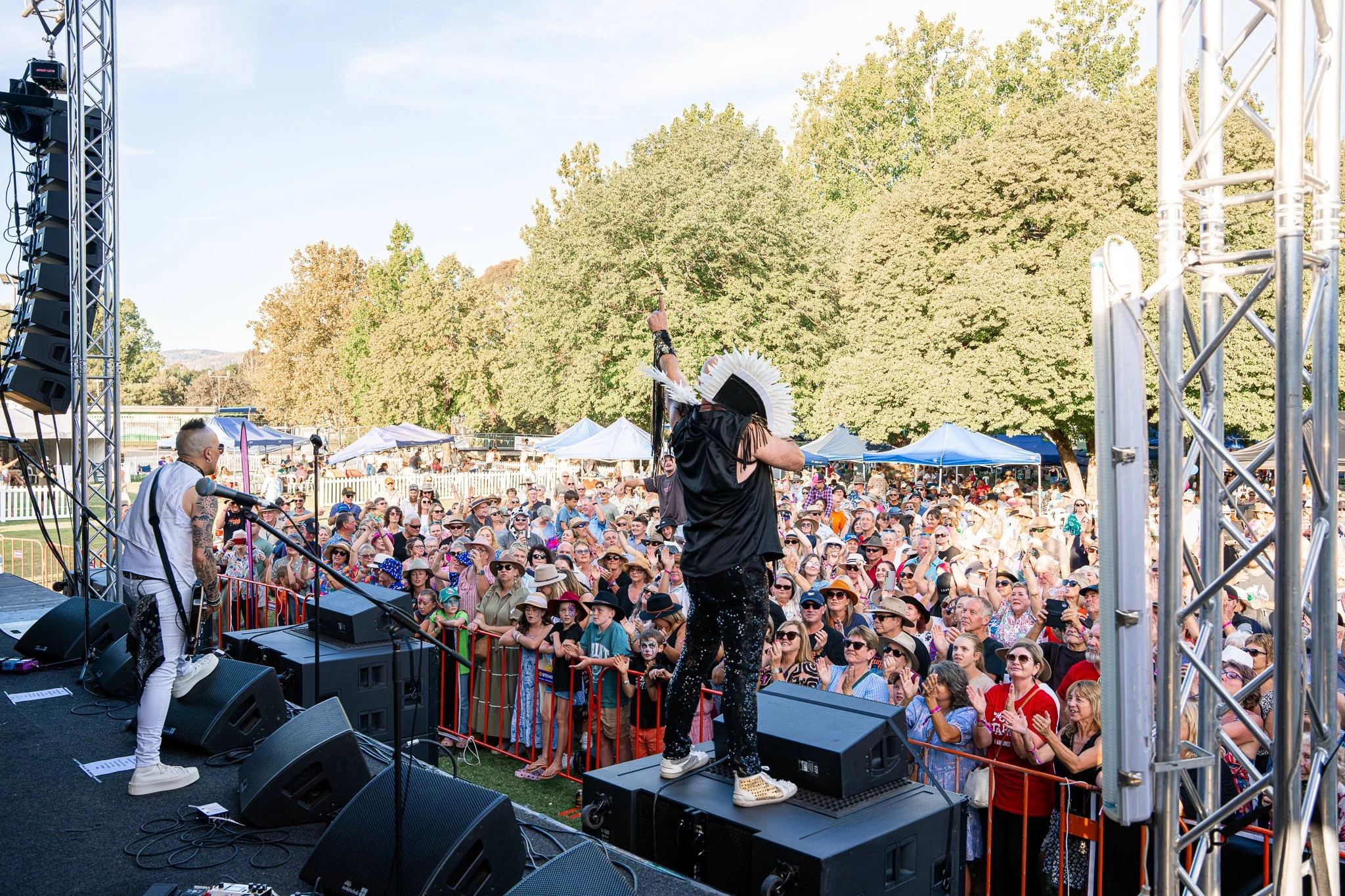 A lively outdoor concert with performers on stage and a large, engaged crowd under a clear sky, surrounded by trees.