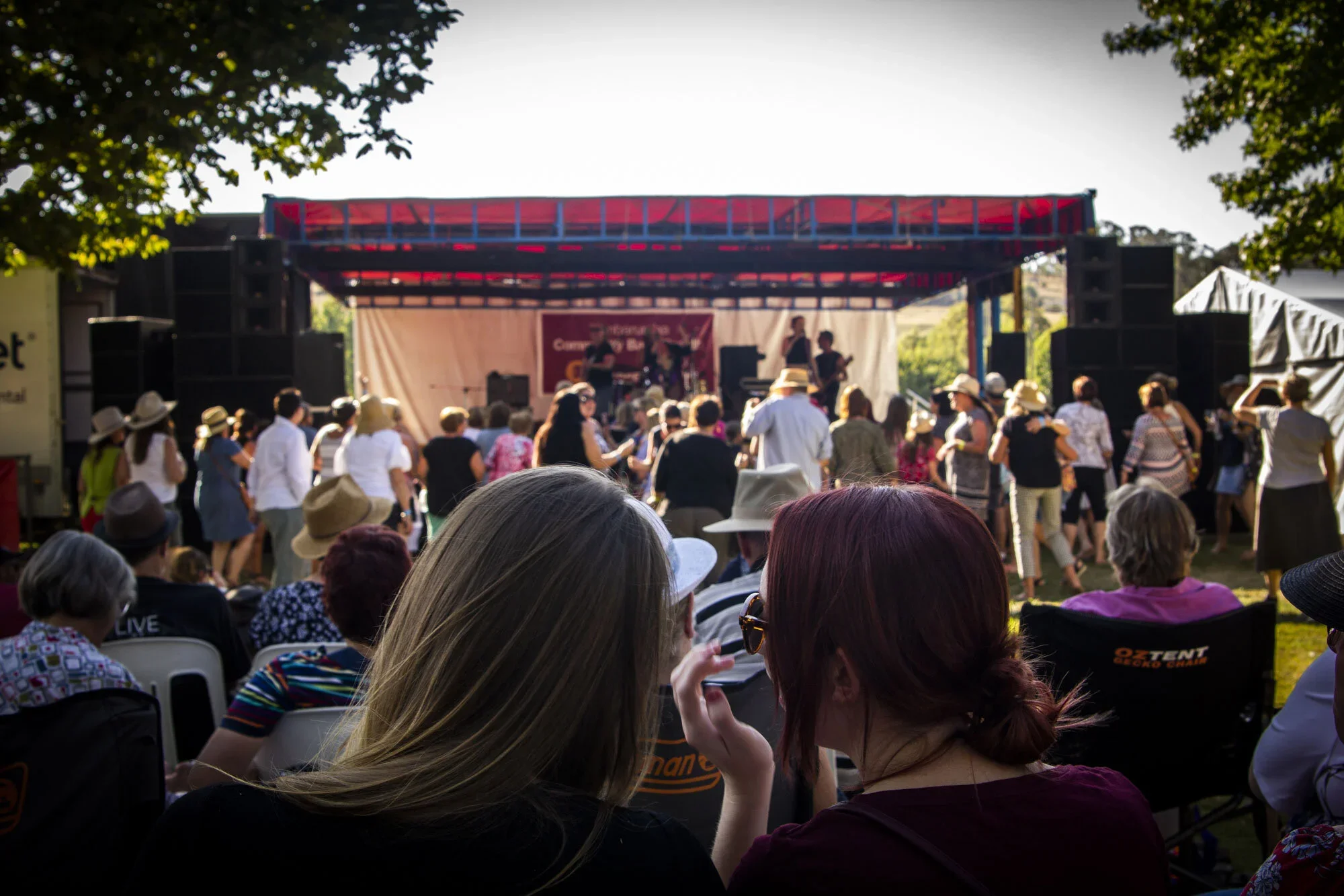 People gathered outdoors at a concert or event with a band performing on a stage, many wearing hats and sitting or standing. The stage is set under trees with sunlight filtering through the leaves.