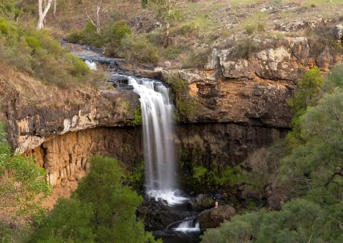 A waterfall flowing over a rocky cliff into a small pool surrounded by green bushes and trees.
