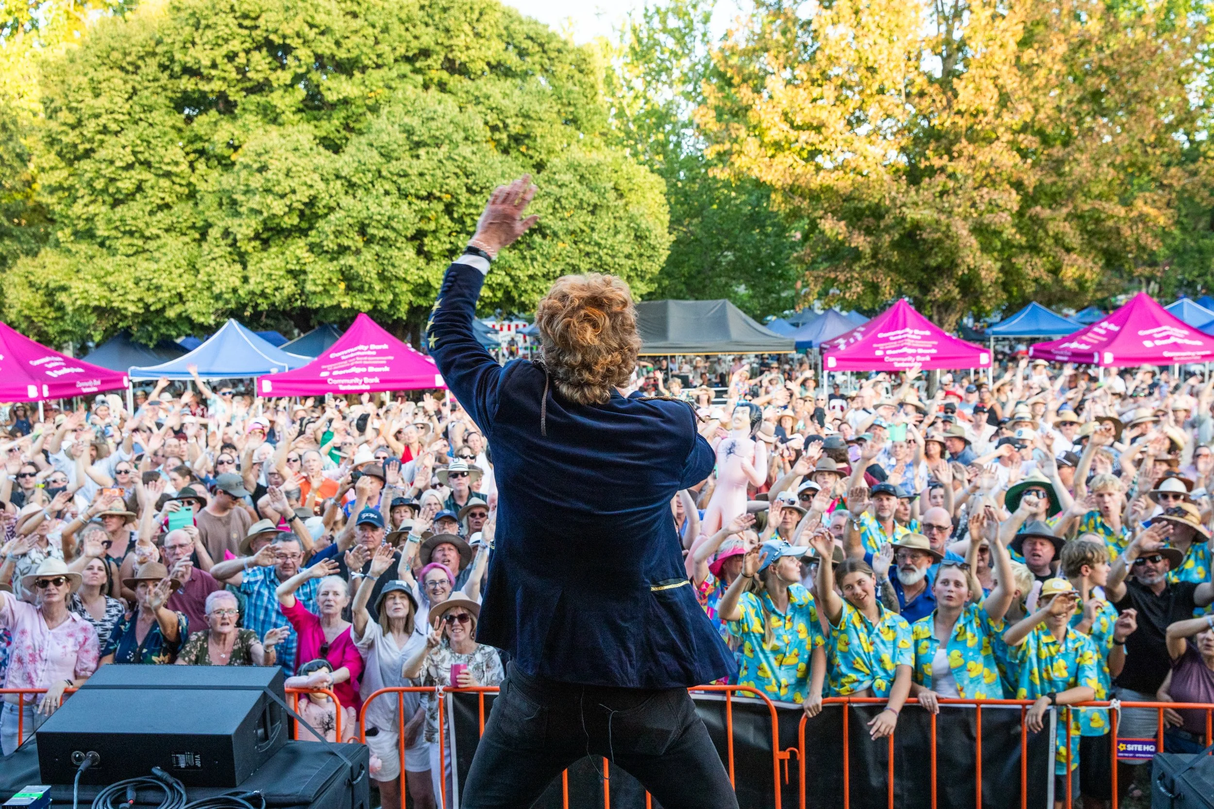 A woman leading a large outdoor crowd event with people wearing hats and colorful clothing, surrounded by green trees, and pink tents in the background.