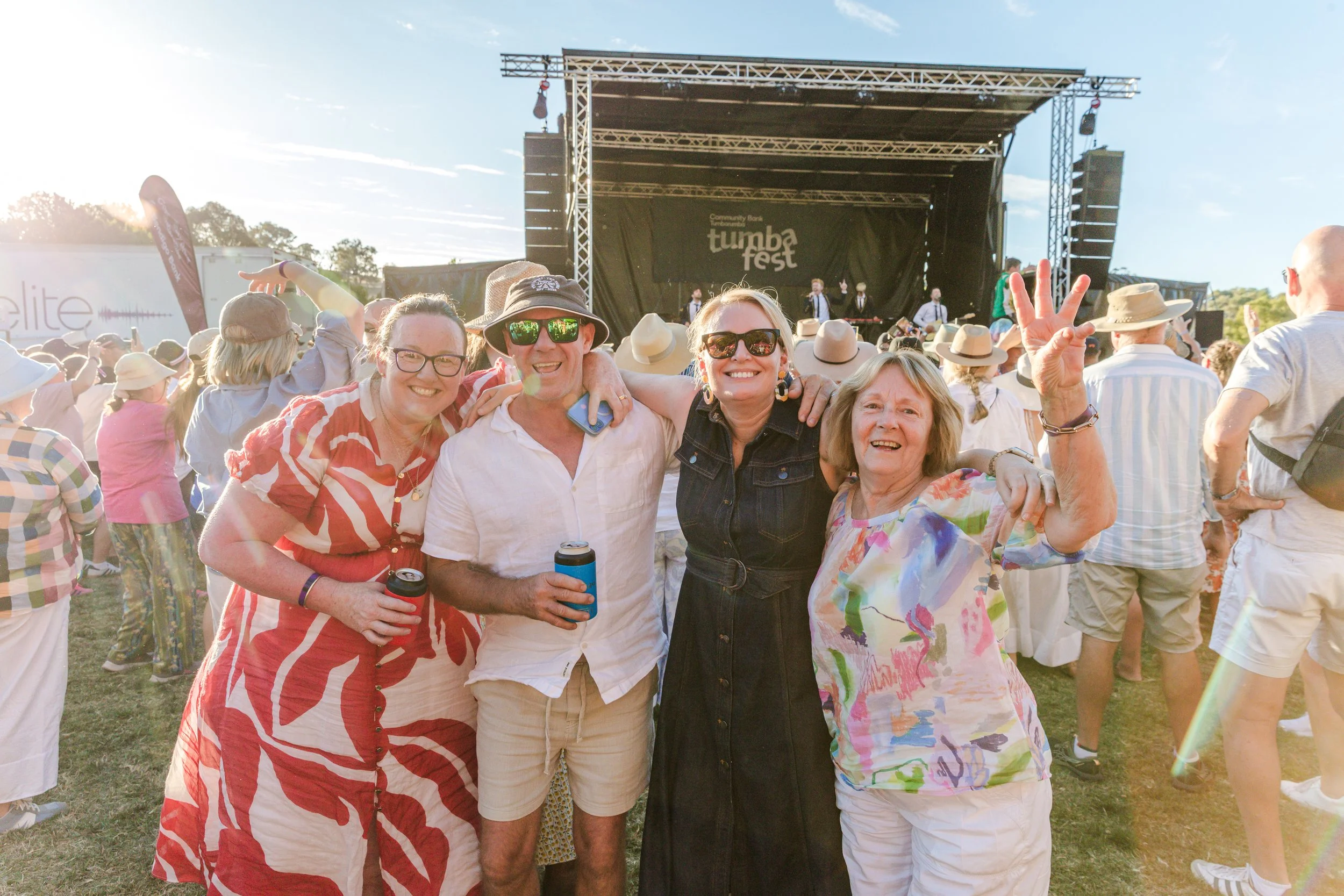 People enjoying a music festival outdoors with a stage in the background, some wearing sunglasses and summer clothing, sunny weather, and a crowd gathered around.