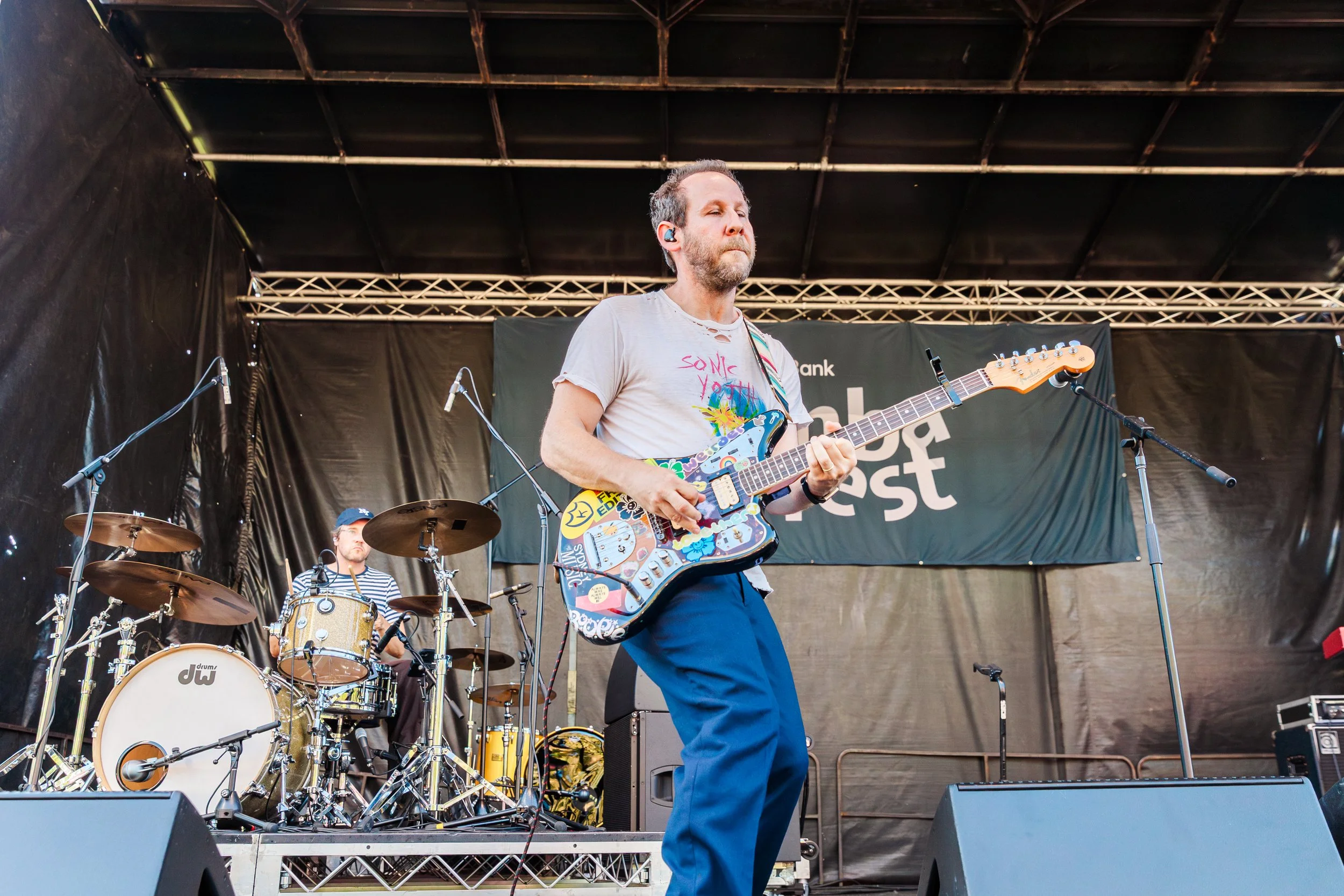 A man playing guitar on stage during a live music performance, with a drum set and band members in the background.