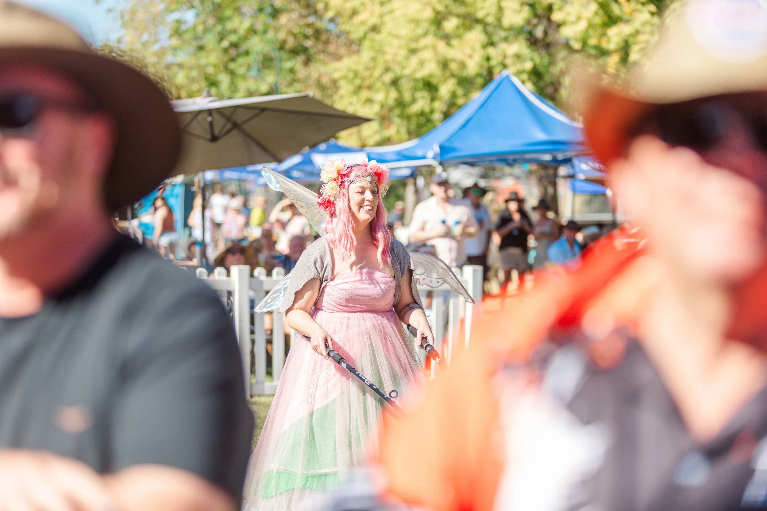 A woman dressed as a fairy with pink hair, wings, a flower crown, and a pink dress, smiling while walking outdoors at a festival or fair, with booths and people in the background.