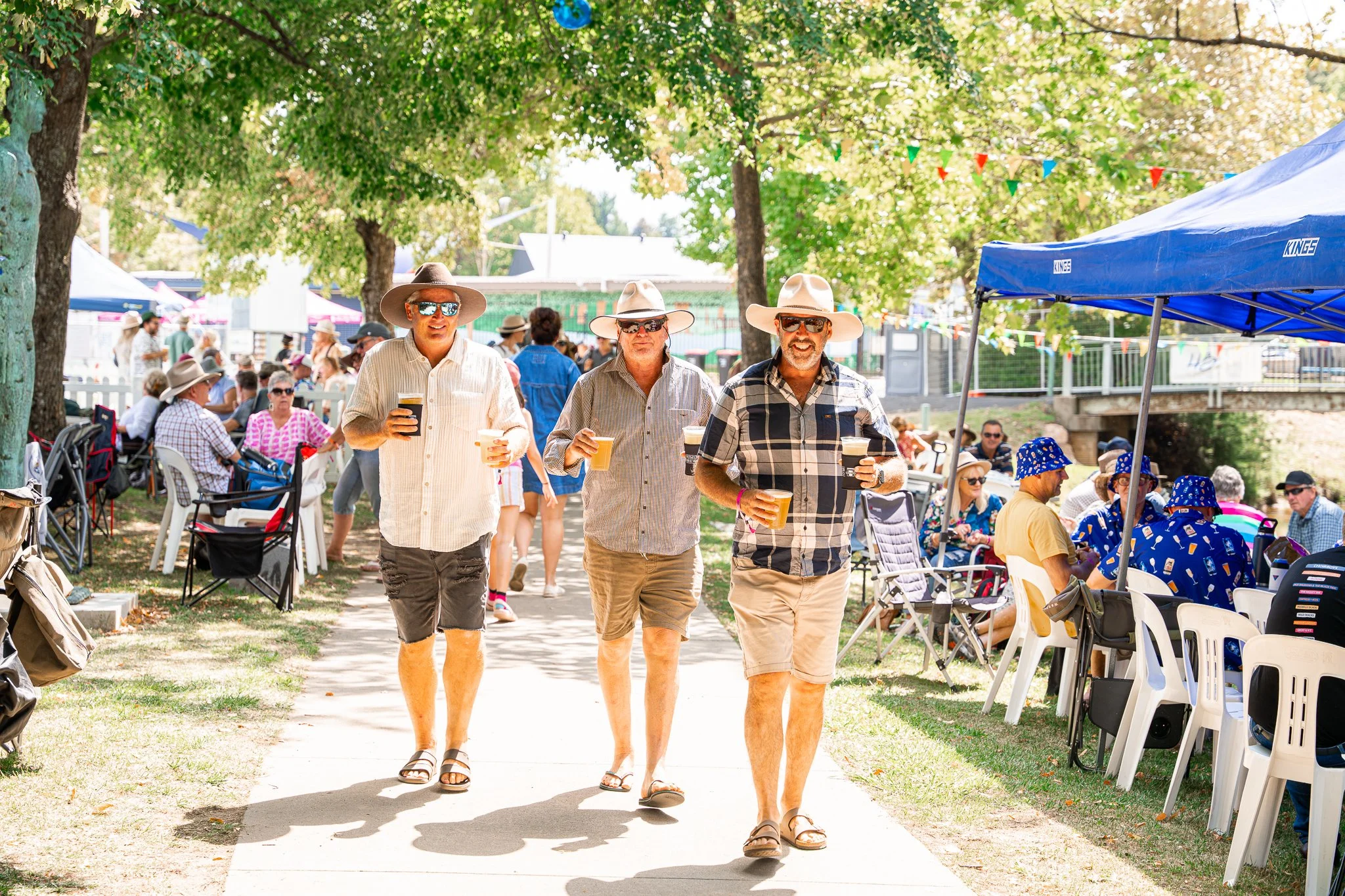 Three men walking on a park pathway at an outdoor event, holding drinks, wearing casual summer clothes, hats, and sunglasses, with many people sitting and standing under trees and tents around them.