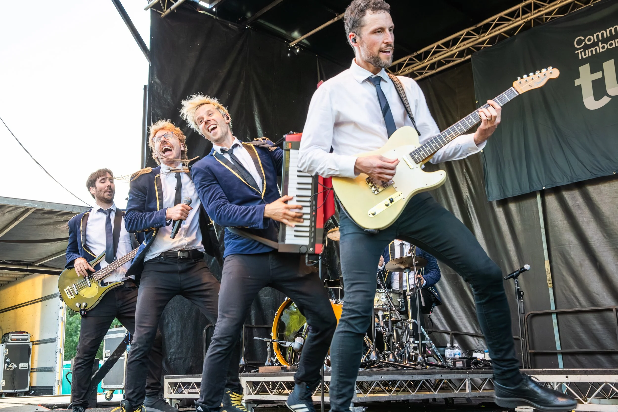 Band of five male musicians performing on an outdoor stage, playing guitars, keyboard, and drums, dressed in formal attire with some wearing jackets and ties, during daylight.