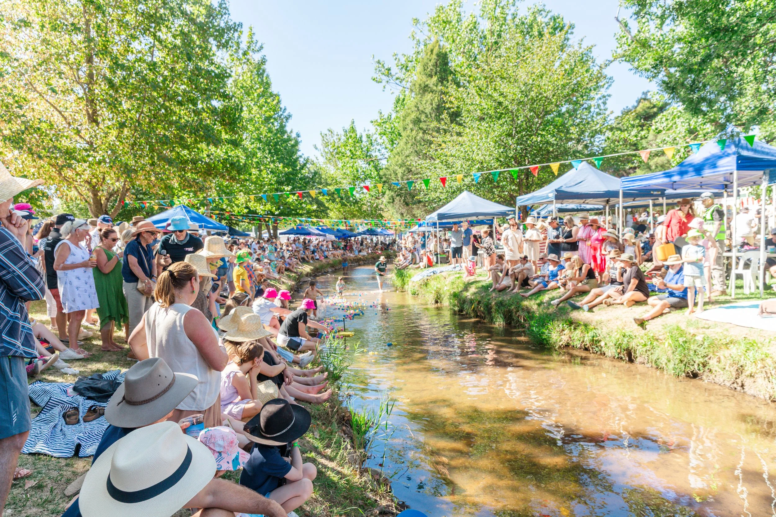 Crowd of people sitting and watching a small stream or river, decorated with colorful bunting flags and surrounded by green trees on a sunny day.