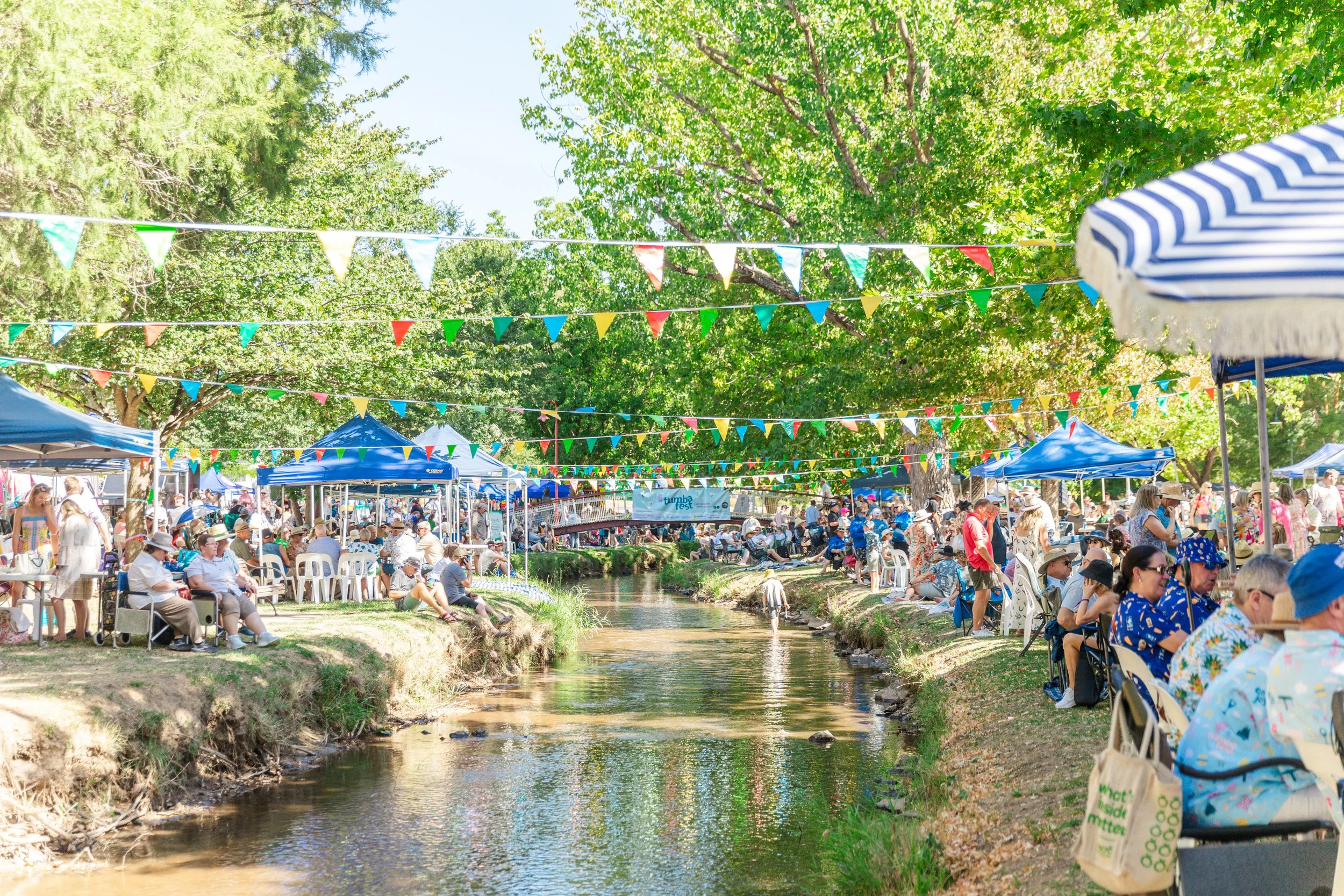 A lively outdoor event with many people sitting on chairs beside a small creek, under trees decorated with colorful bunting flags, with tents and a clear sky.