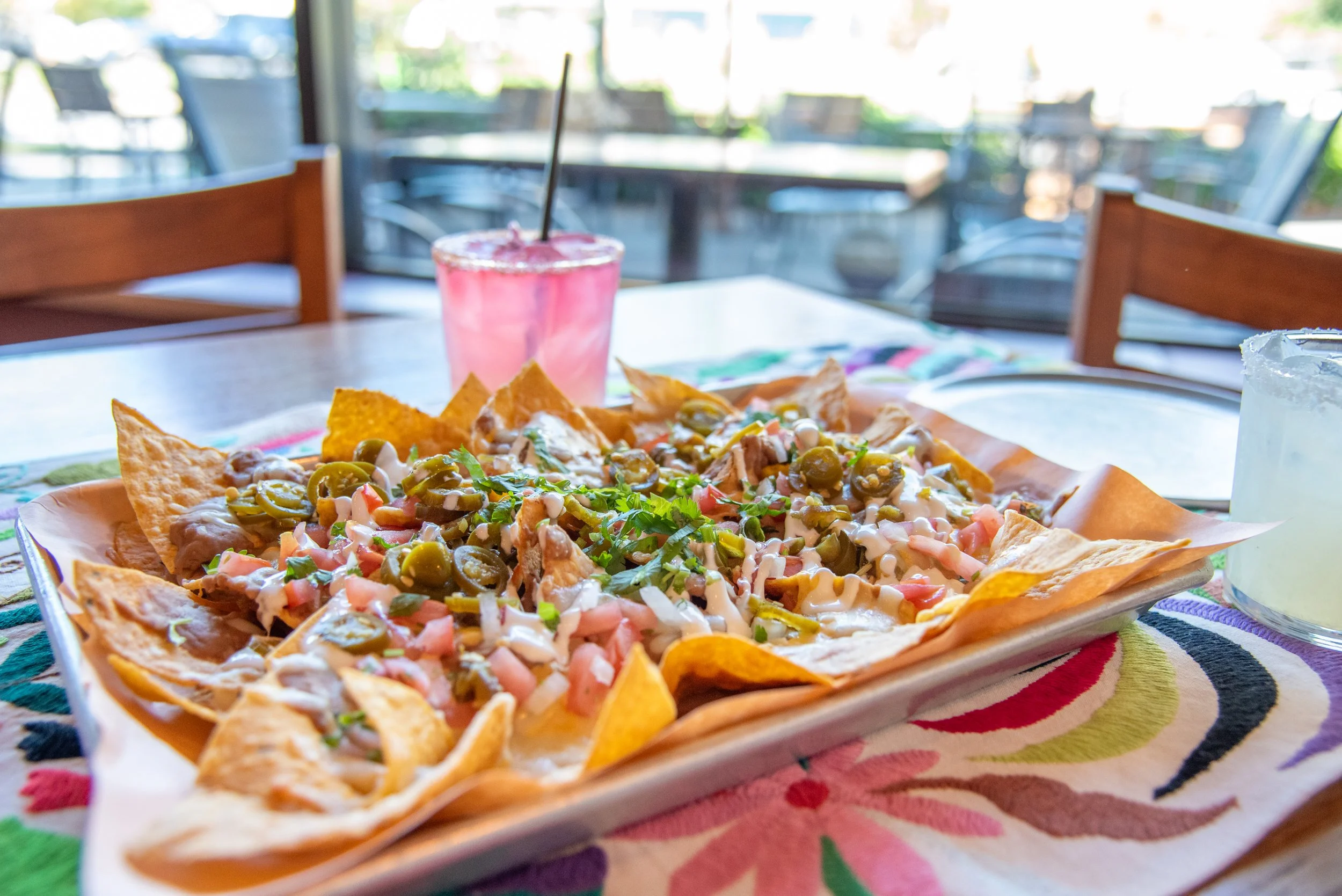 A plate of nachos topped with cheese, jalapeños, tomatoes, and cilantro, placed on a colorful striped tablecloth with a pink drink in the background and a blurred indoor restaurant setting.