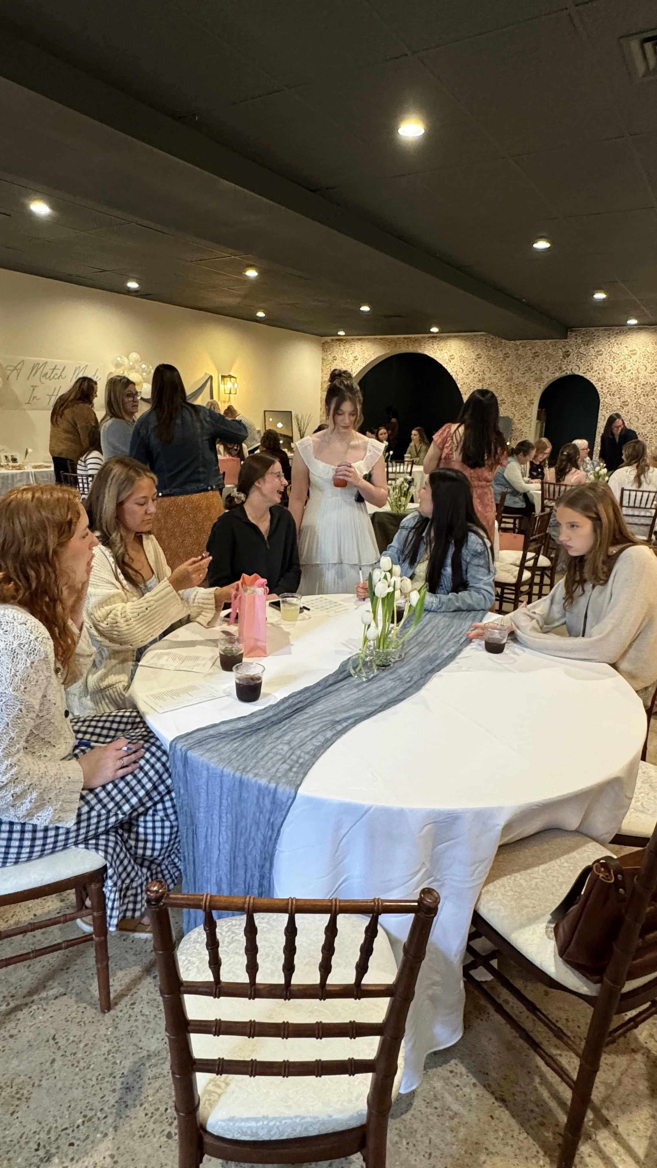 Group of women sitting around a large round table with drinks, floral centerpiece, and event programs, at a social gathering or celebration in a decorated room.
