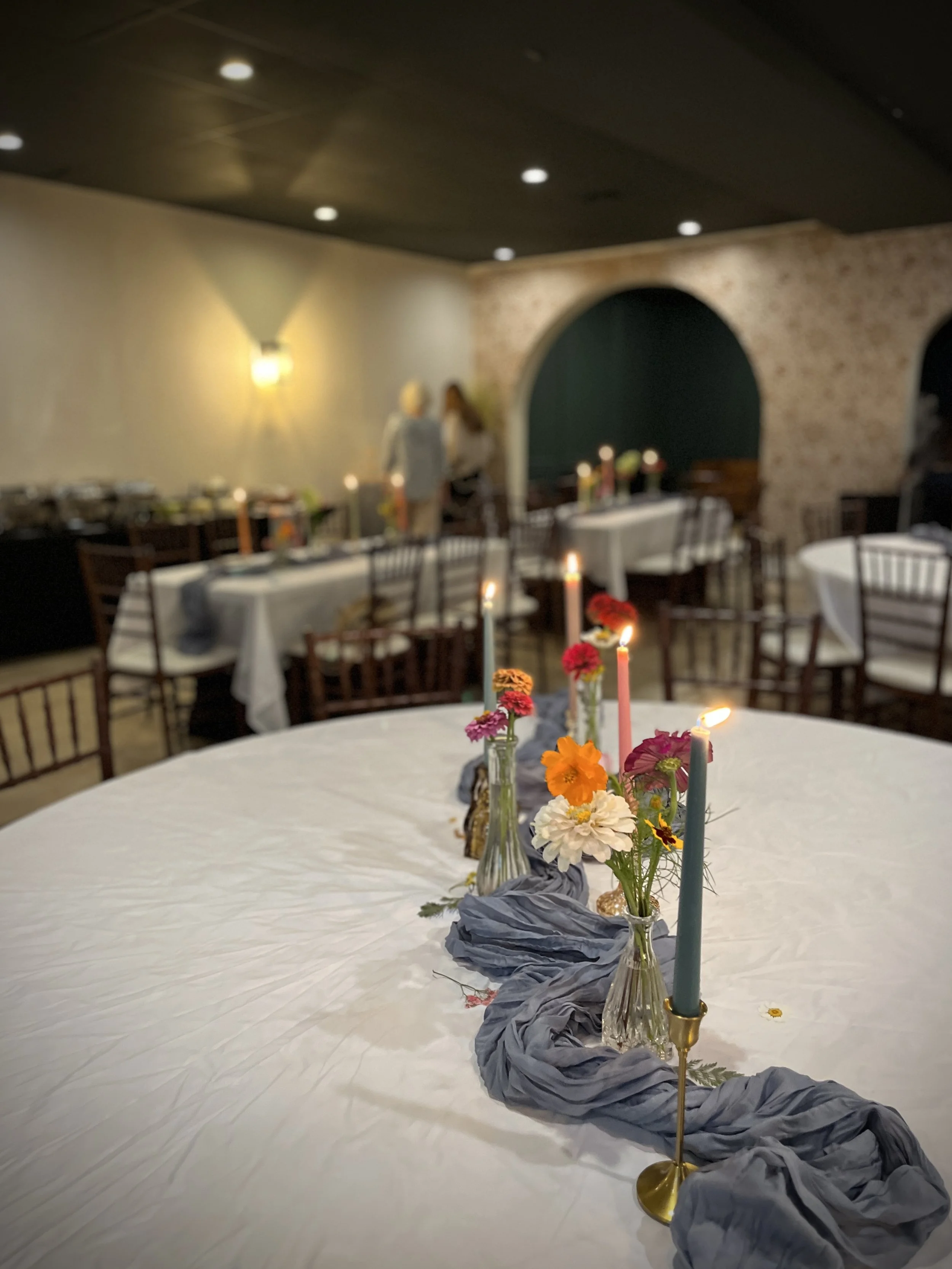 A decorated dining table with flowers and candles, set in an event space with chairs and tables in the background.