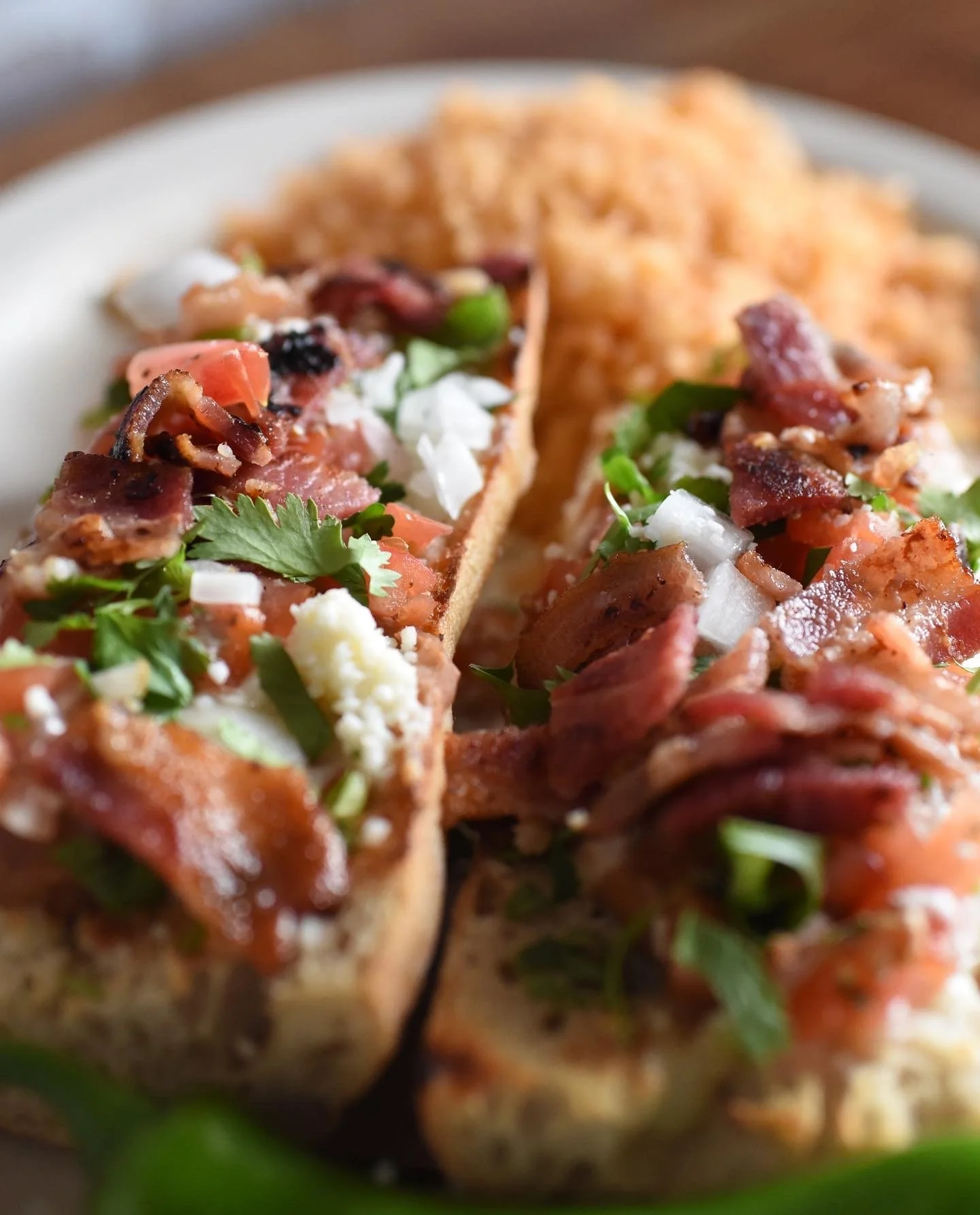 Close-up of a taco with ground meat, diced tomatoes, chopped onions, shredded cheese, cilantro, and bacon, with a side of orange rice in the background.