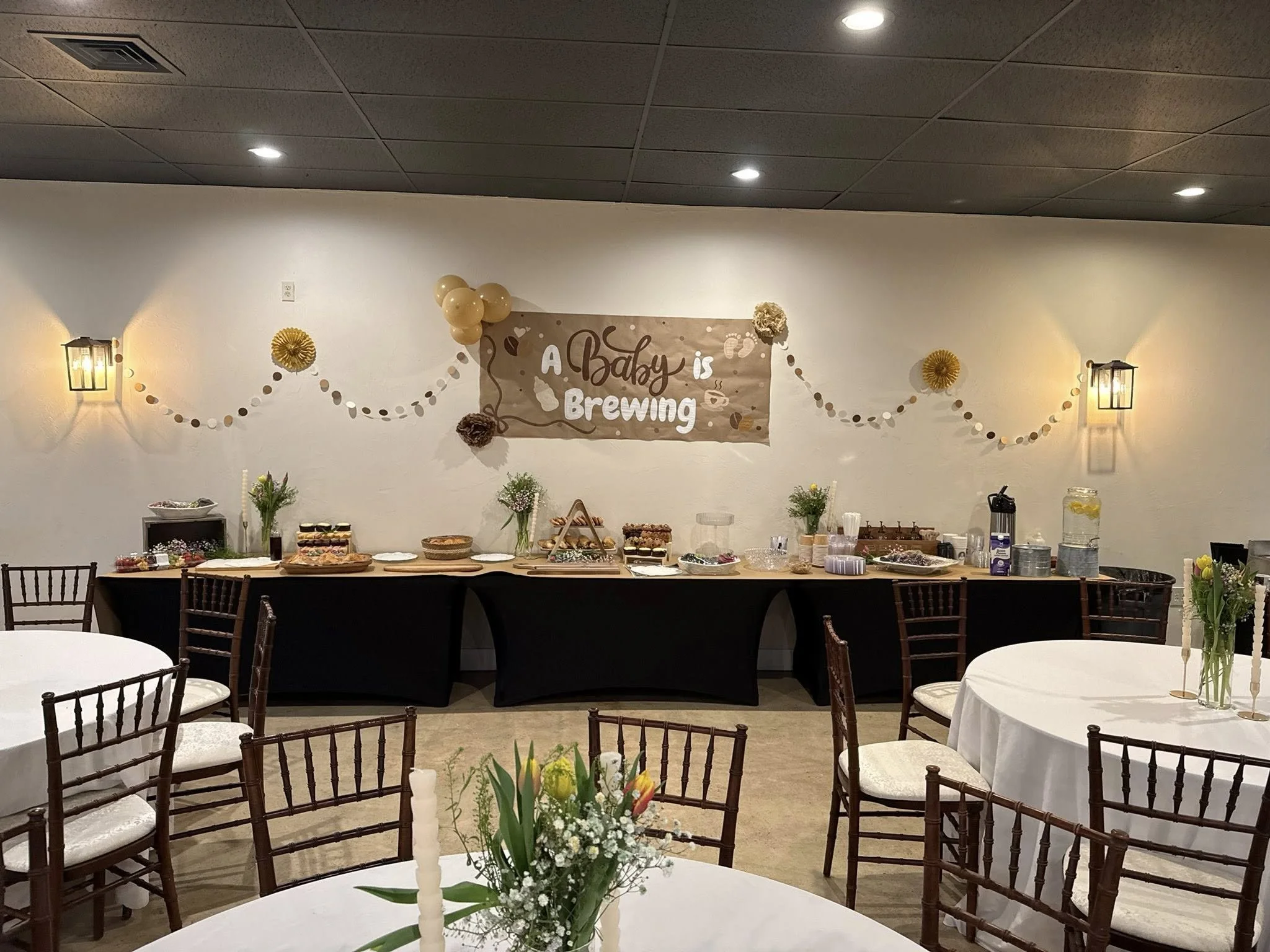 Wedding reception with a buffet table, floral centerpieces, and a banner that reads 'A Baby is Brewing' decorated with balloons and paper flowers.
