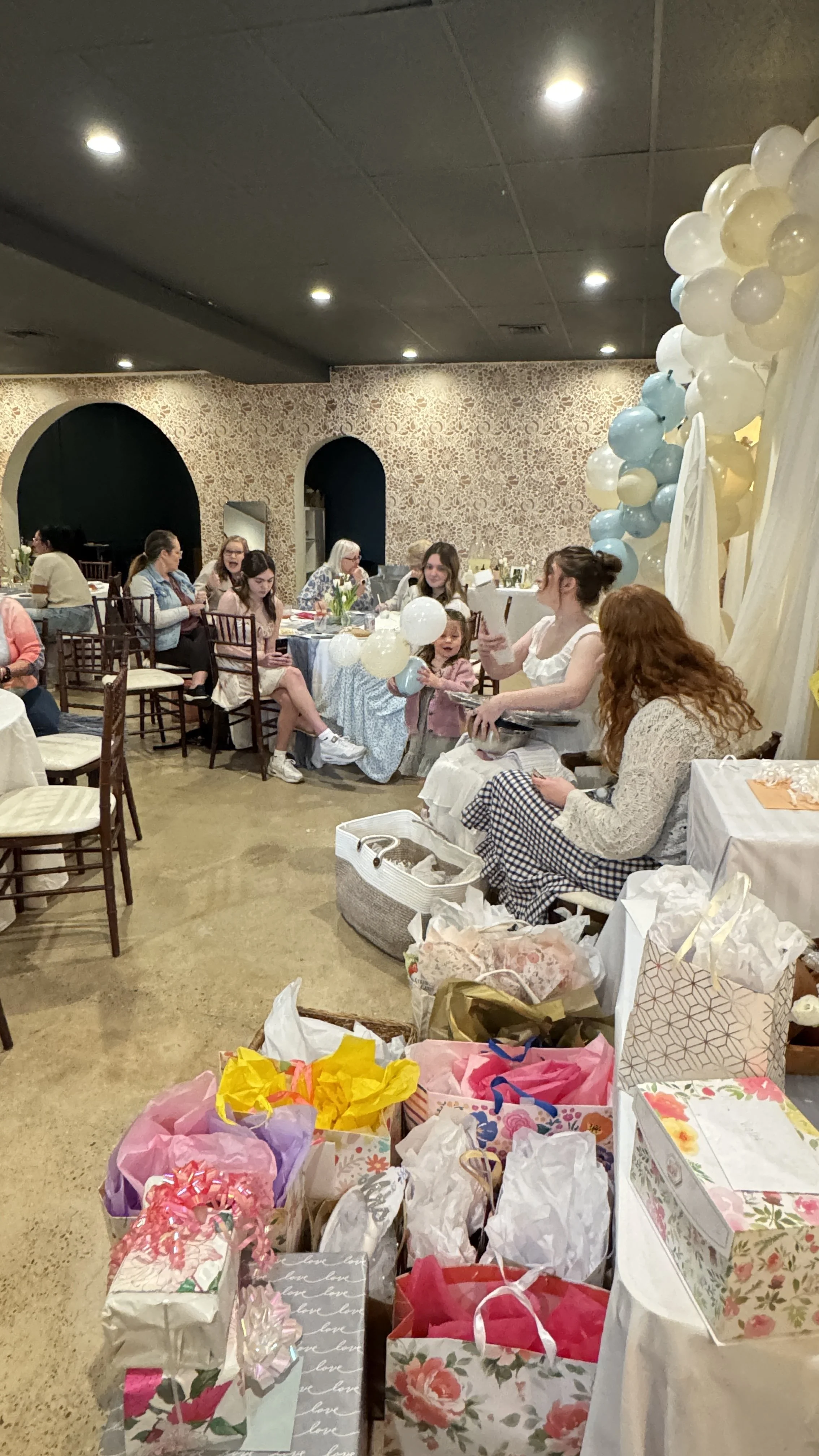 A gathering or celebration with people sitting around tables, decorated with balloons, flowers, and gift bags, indicating an event like a birthday or shower.
