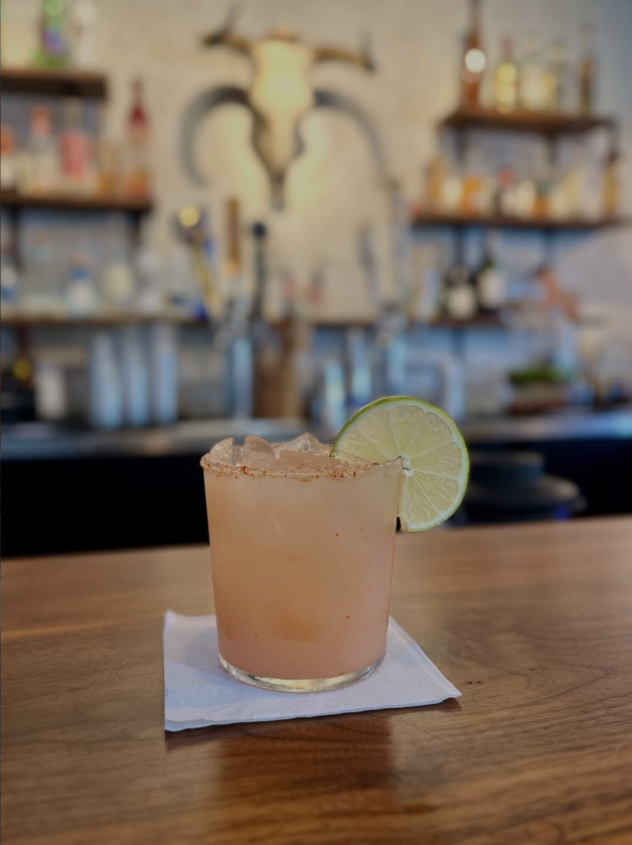 A pink cocktail garnished with a lime wedge, served in a short glass on a white napkin on a wooden table, with a blurred bar background.