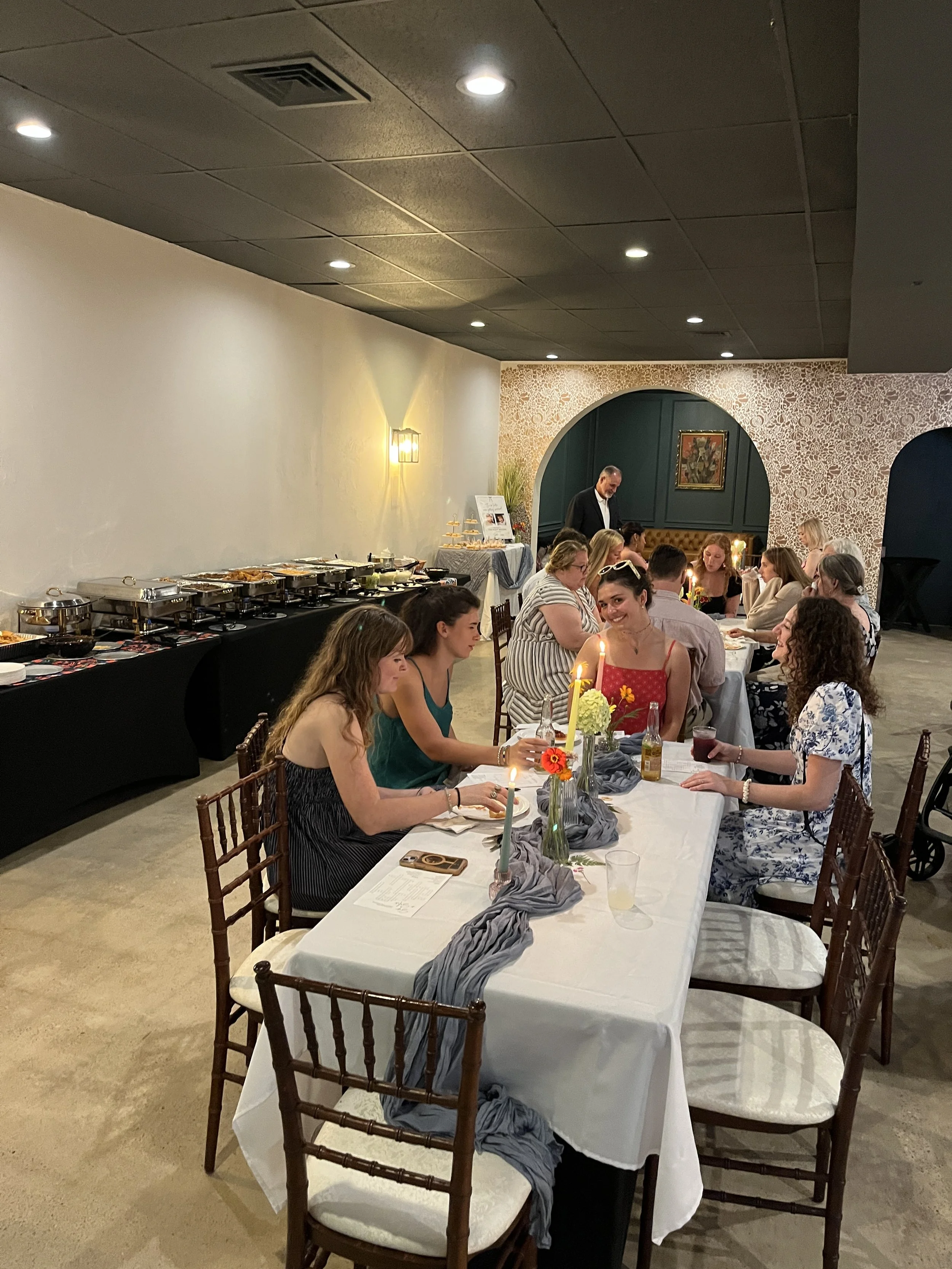 People dining at a restaurant with decorated tables, candles, and floral centerpieces, while a buffet with food is set up on the side. The background features a patterned wall and a man in a suit standing near a table.