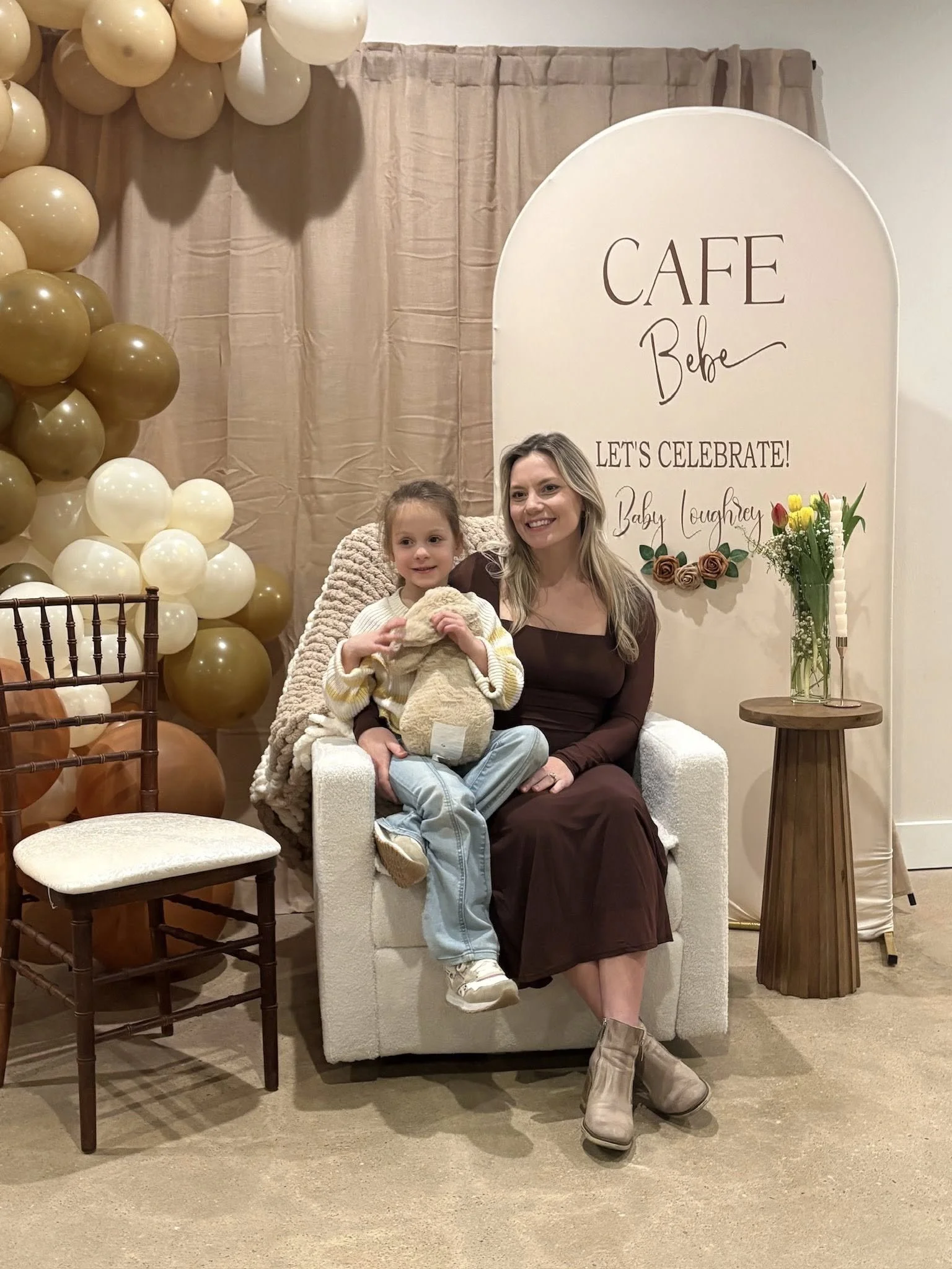 A woman and a young girl sitting on a white armchair, smiling. Behind them is a beige backdrop with a banner that reads "Café Bebe," and balloons in shades of gold and cream are on the left side. There is a small round wooden table with a tall glass vase of yellow and red flowers on the right.