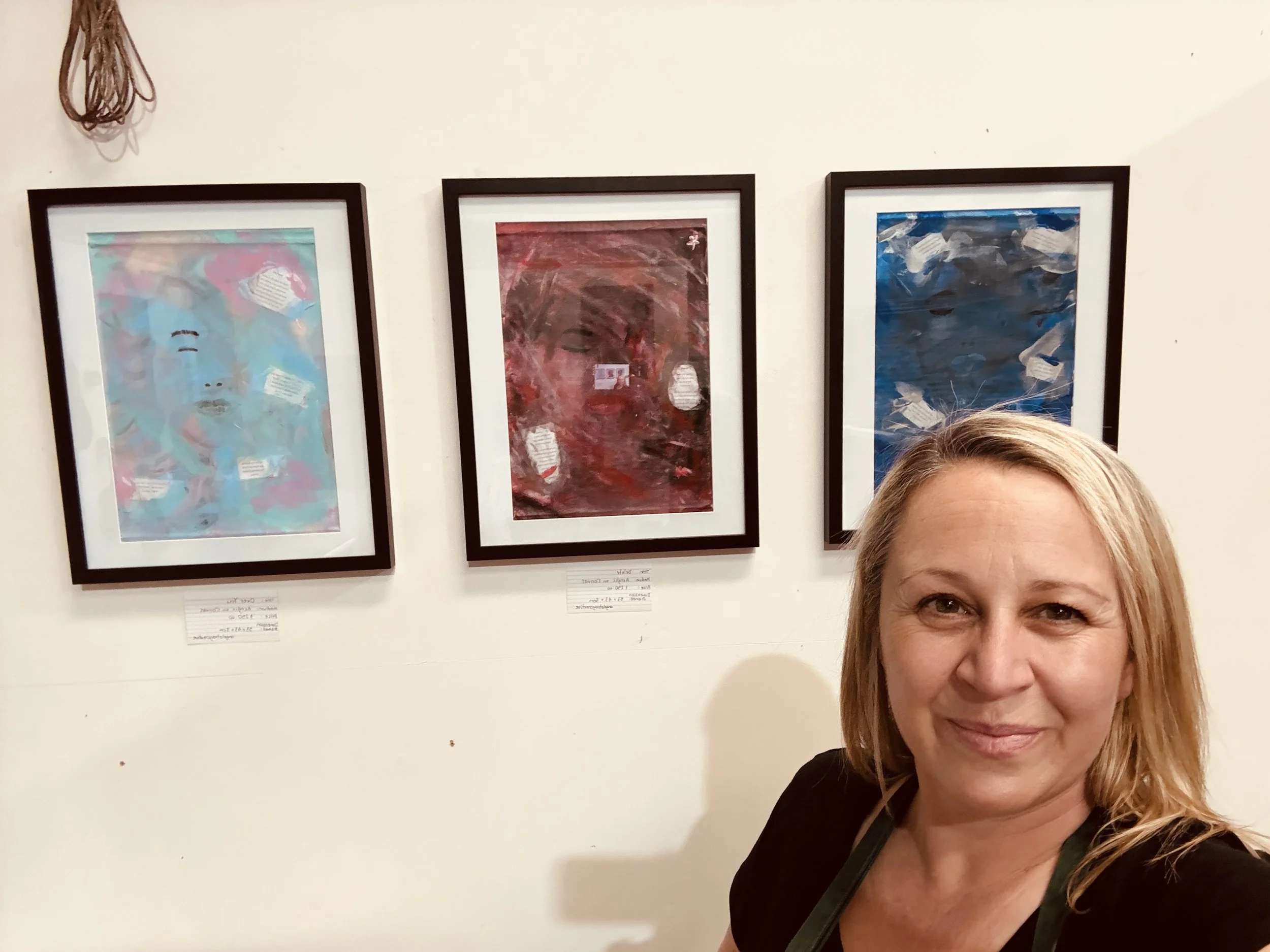 A woman with blonde hair smiling in front of three framed abstract art paintings on a white wall.