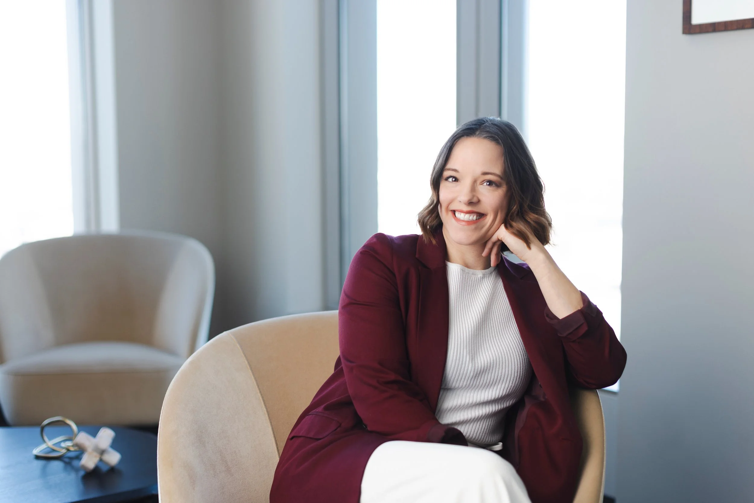 A woman with shoulder-length brown hair, smiling, wearing a white top and a burgundy blazer, sitting at a table in a bright room with large windows.