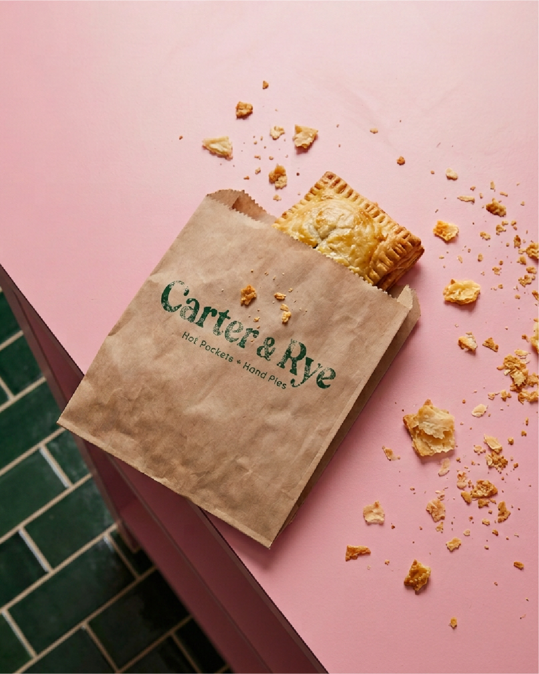 A pastry partially inside a brown paper bag on a pink surface, with crumbs scattered around.