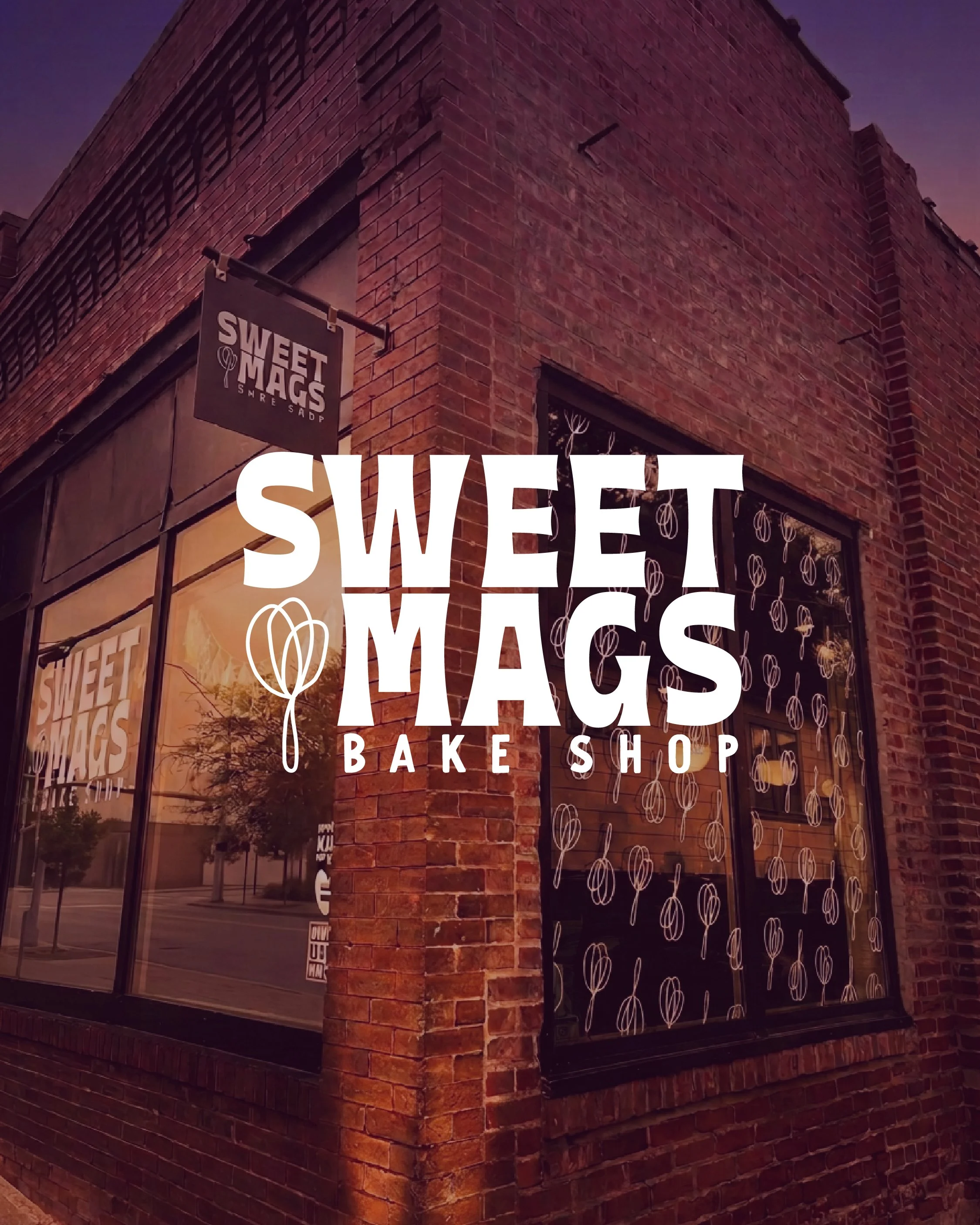 A brick building with large windows display signs for 'Sweet Mags Bake Shop.' The signs feature the shop's logo of a balloon and the words 'Sweet Mags' in bold white lettering.