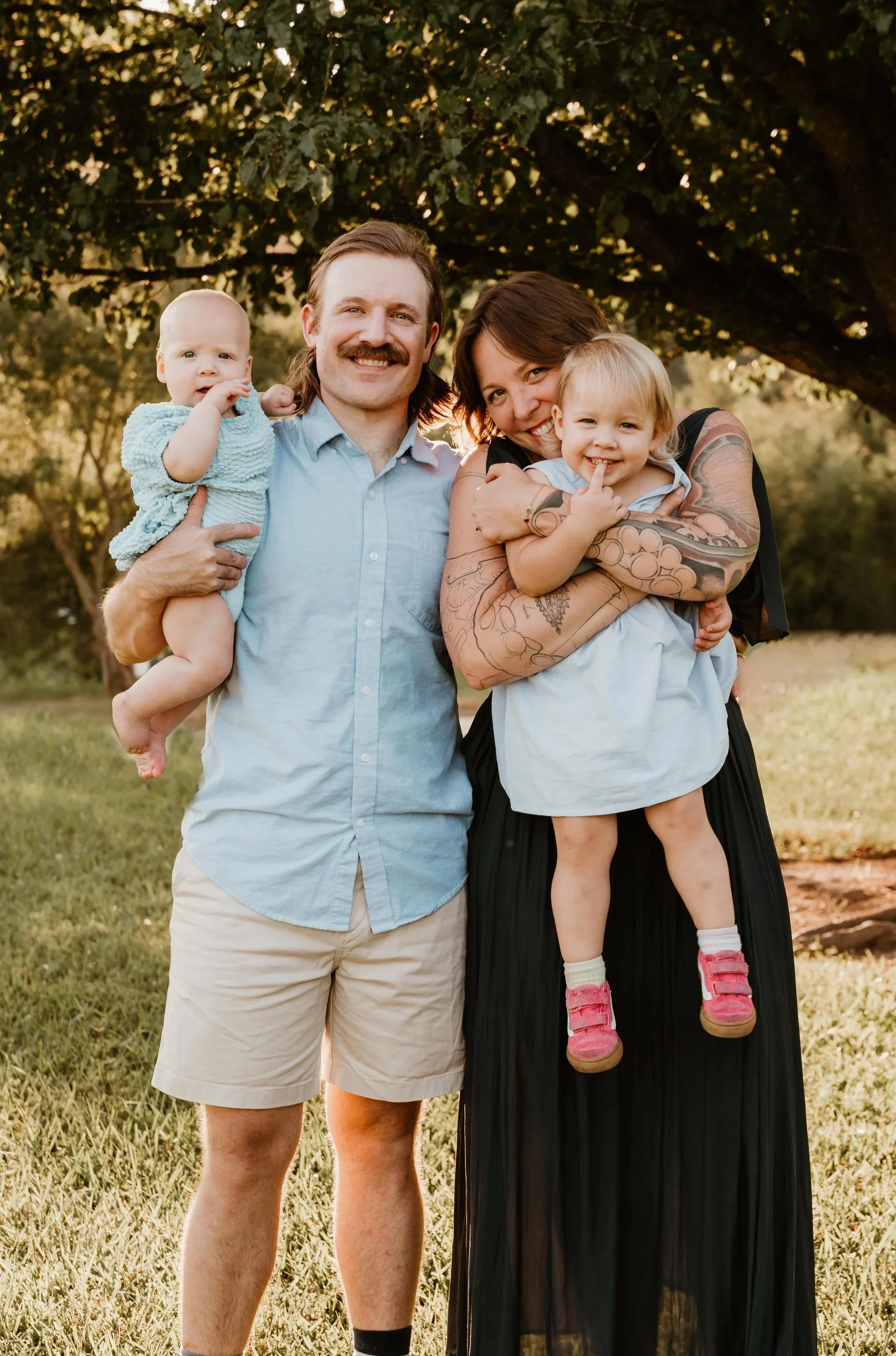 A happy family of four outdoors, standing on grass near a large tree, during sunset. The father, with a mustache, is holding a young child with short blond hair in a blue outfit. The mother, with tattoos on her arms, is holding a smiling toddler with blond hair in a light blue dress.