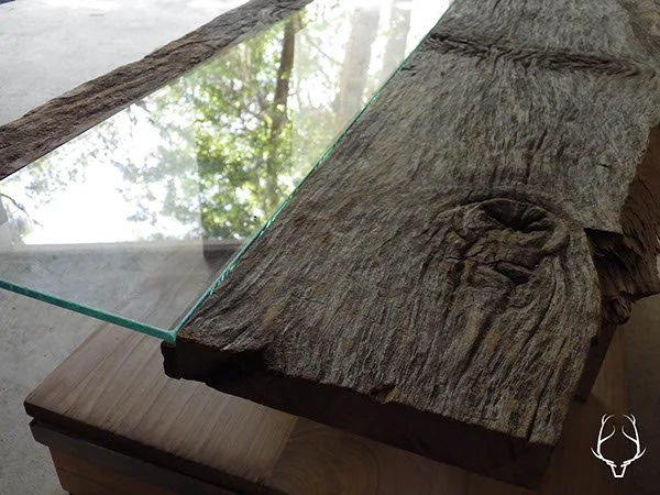Close-up of a piece of weathered wood with a glass table top on top of it, reflecting trees and sky from the window above.