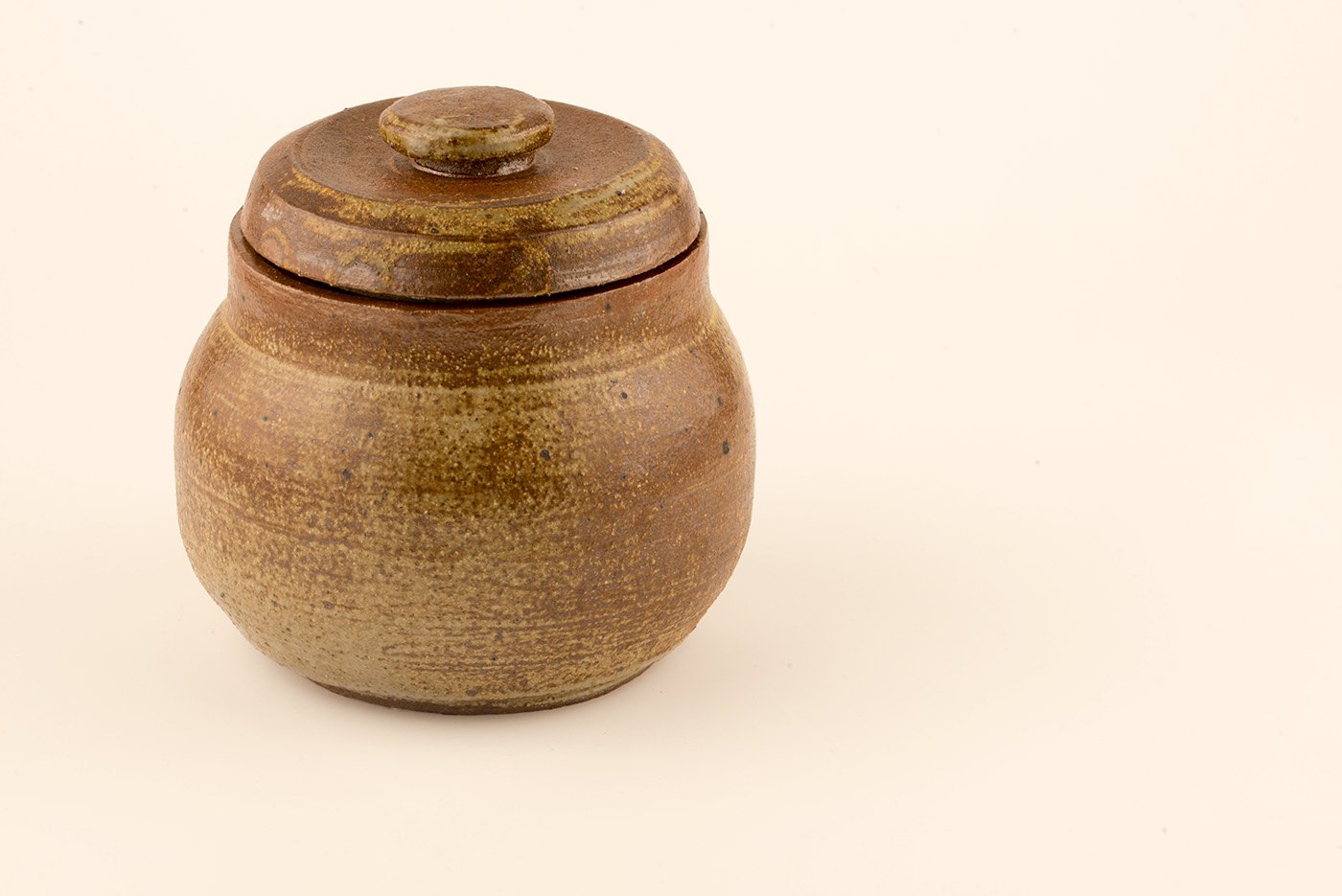 A small, round, brown ceramic jar with a matching lid against a plain light background.