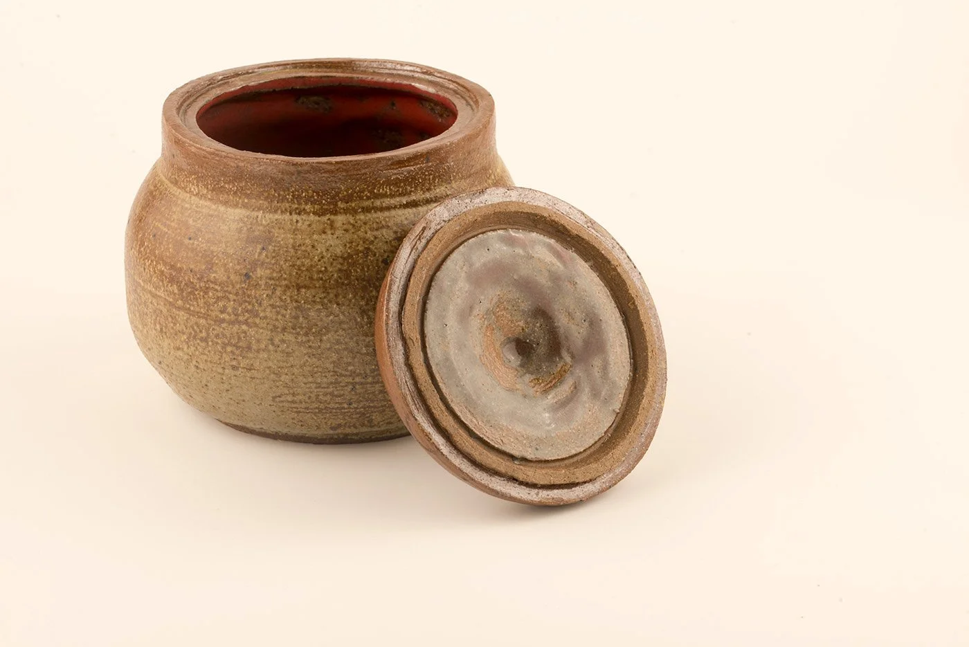 A round, brown ceramic jar with an open lid placed beside it, showing the jar's dark interior against a plain, light background.