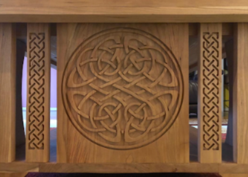 Close-up of a wooden furniture piece with a carved circular Celtic knot design in the center and decorative patterns on the sides.
