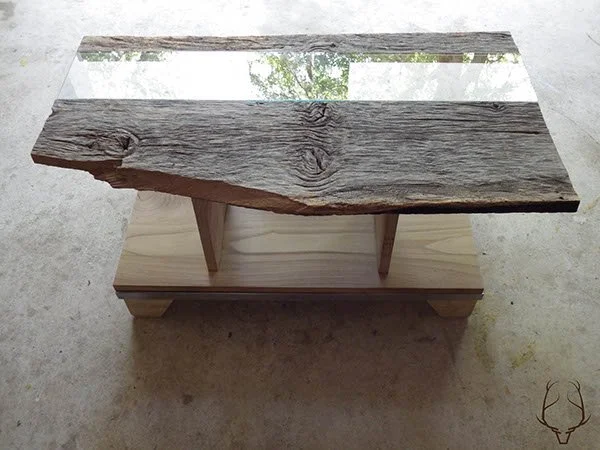 A wooden table with a rustic, weathered wood top, featuring a section of glass or mirror as part of the surface, and supported by a light-colored wooden base with angled legs.
