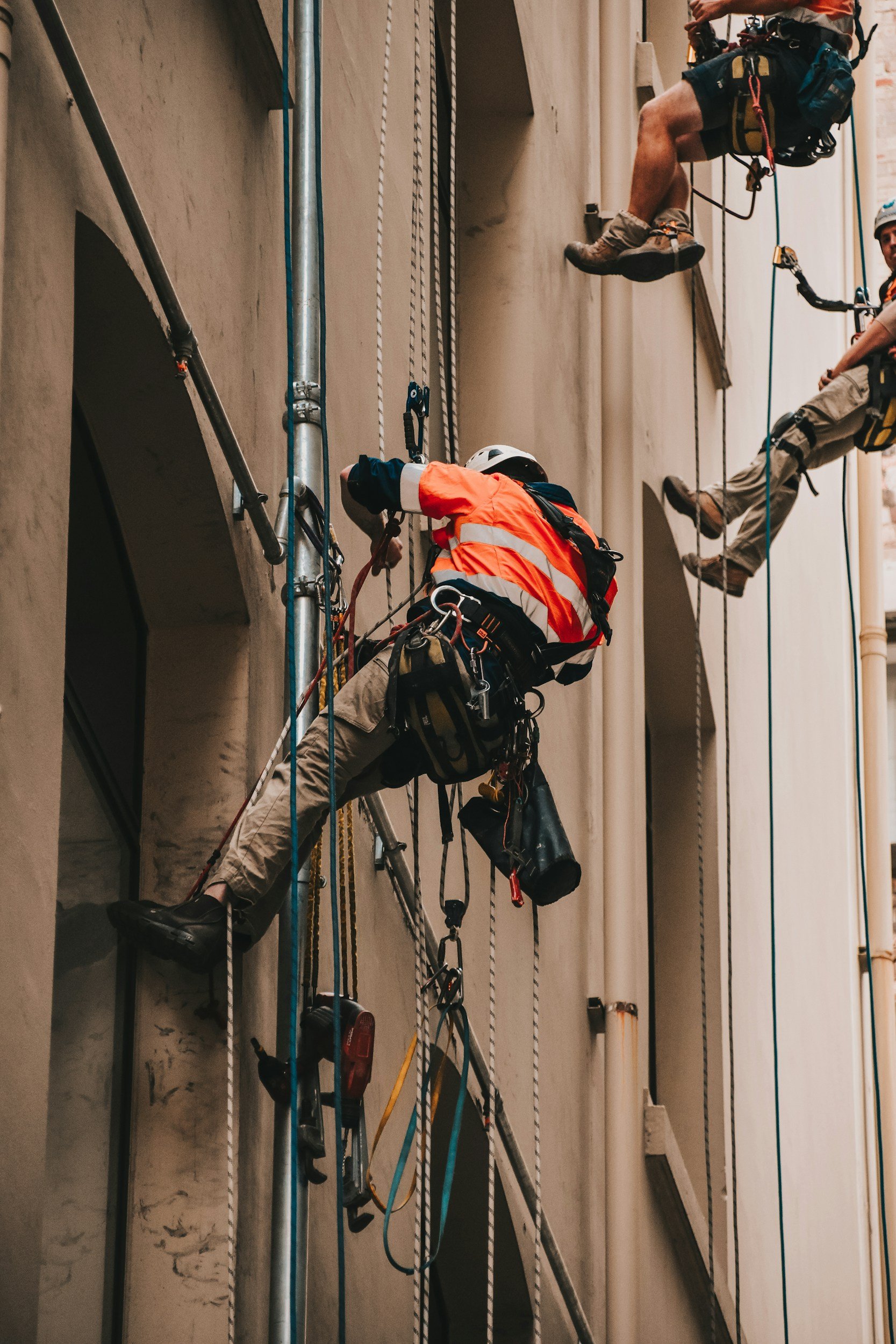 Two workers are rappelling down a building, secured with harnesses and ropes, wearing helmets and safety gear.