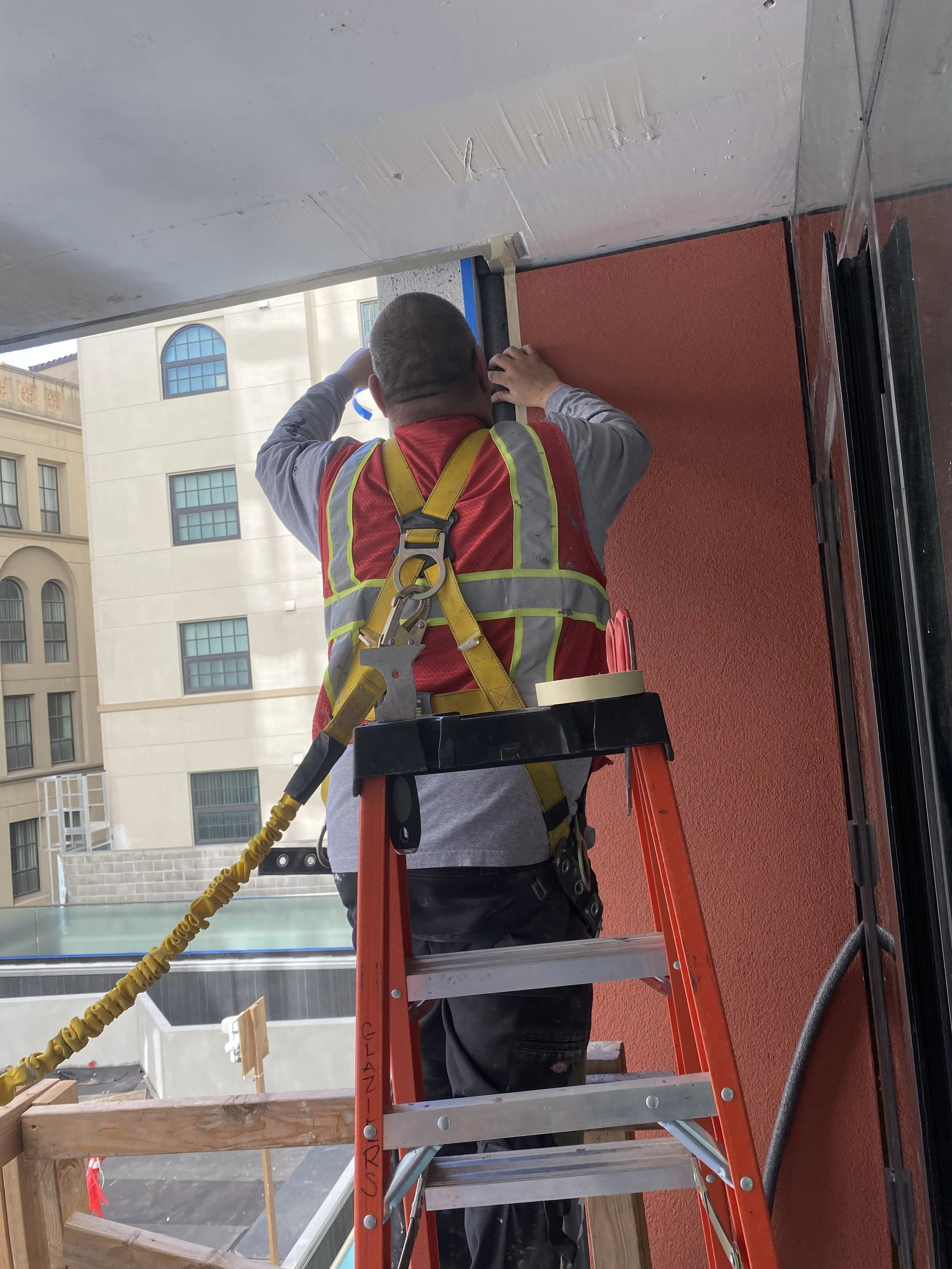 A construction worker with a gray shirt and black pants stands on a red ladder, working on a wall with a cutting tool. The worker is wearing a yellow safety harness and is in an outdoor setting with buildings in the background.