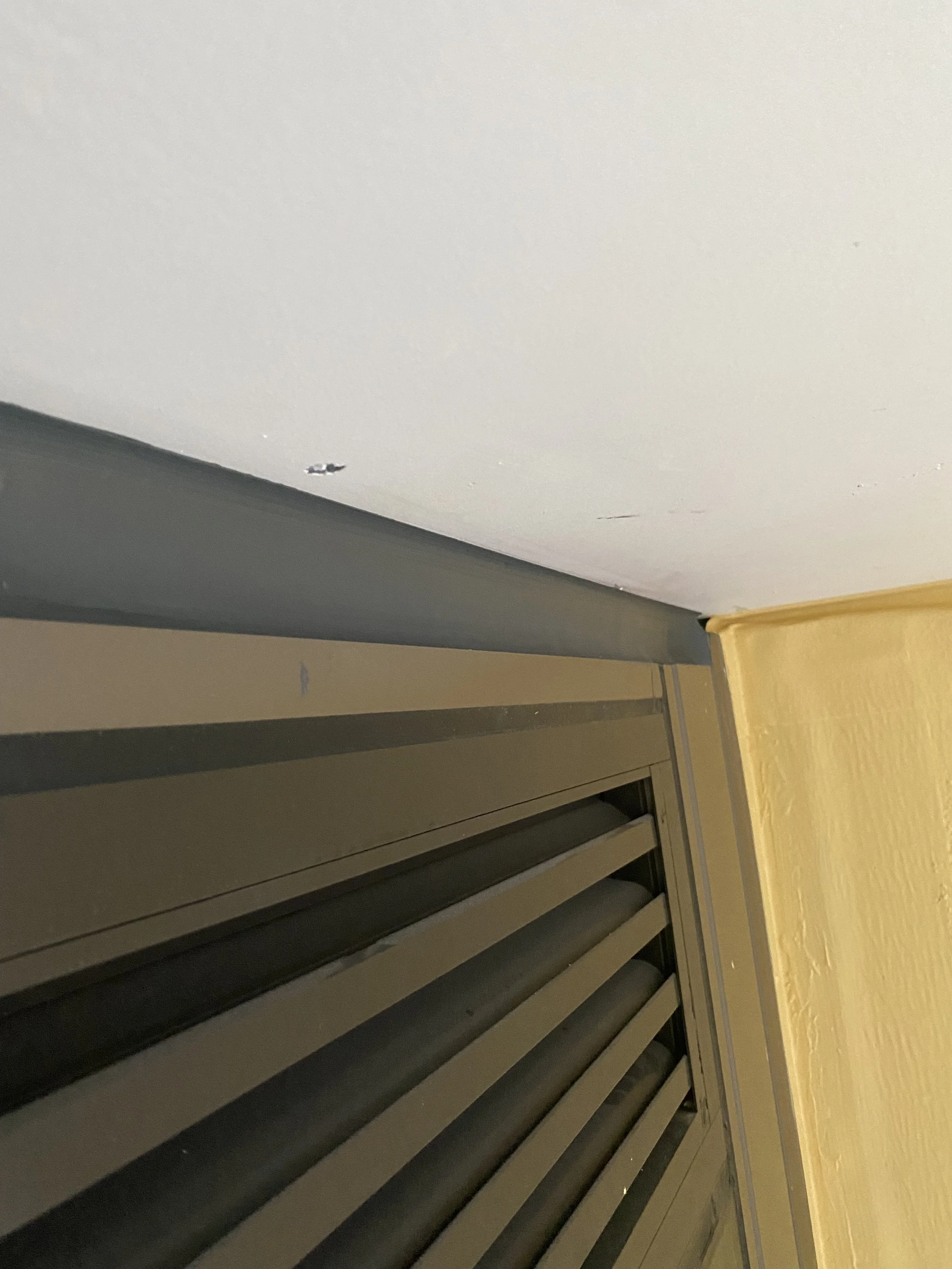 Image shows a view of a dark, metallic, louvered door or vent with black louvers, inside a room with beige walls and a yellow-colored wooden structure or furniture.